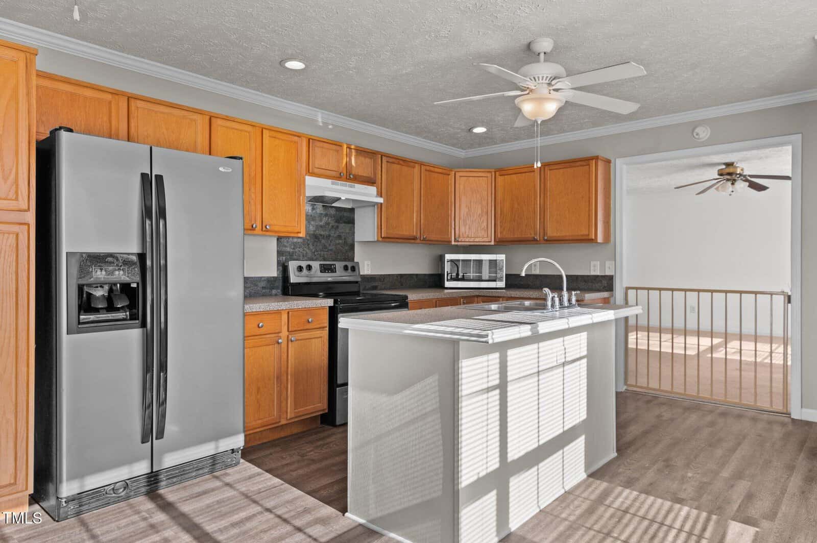 A kitchen with wooden cabinets, a stainless steel refrigerator, and a black stove. An island with a sink sits in the center, and a ceiling fan is above. Light streams in through a window, illuminating the hardwood floor.