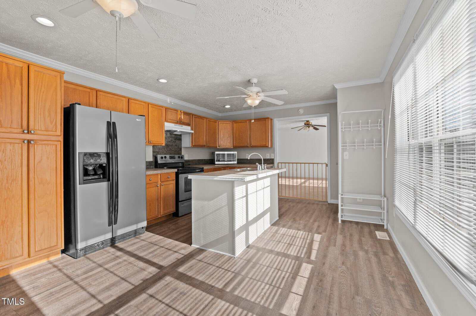 A kitchen with wooden cabinets and a dark countertop. There is a stainless steel refrigerator, an electric stove, and a microwave. An island with a sink is in the center. The flooring is wooden, and large windows bring in natural light.