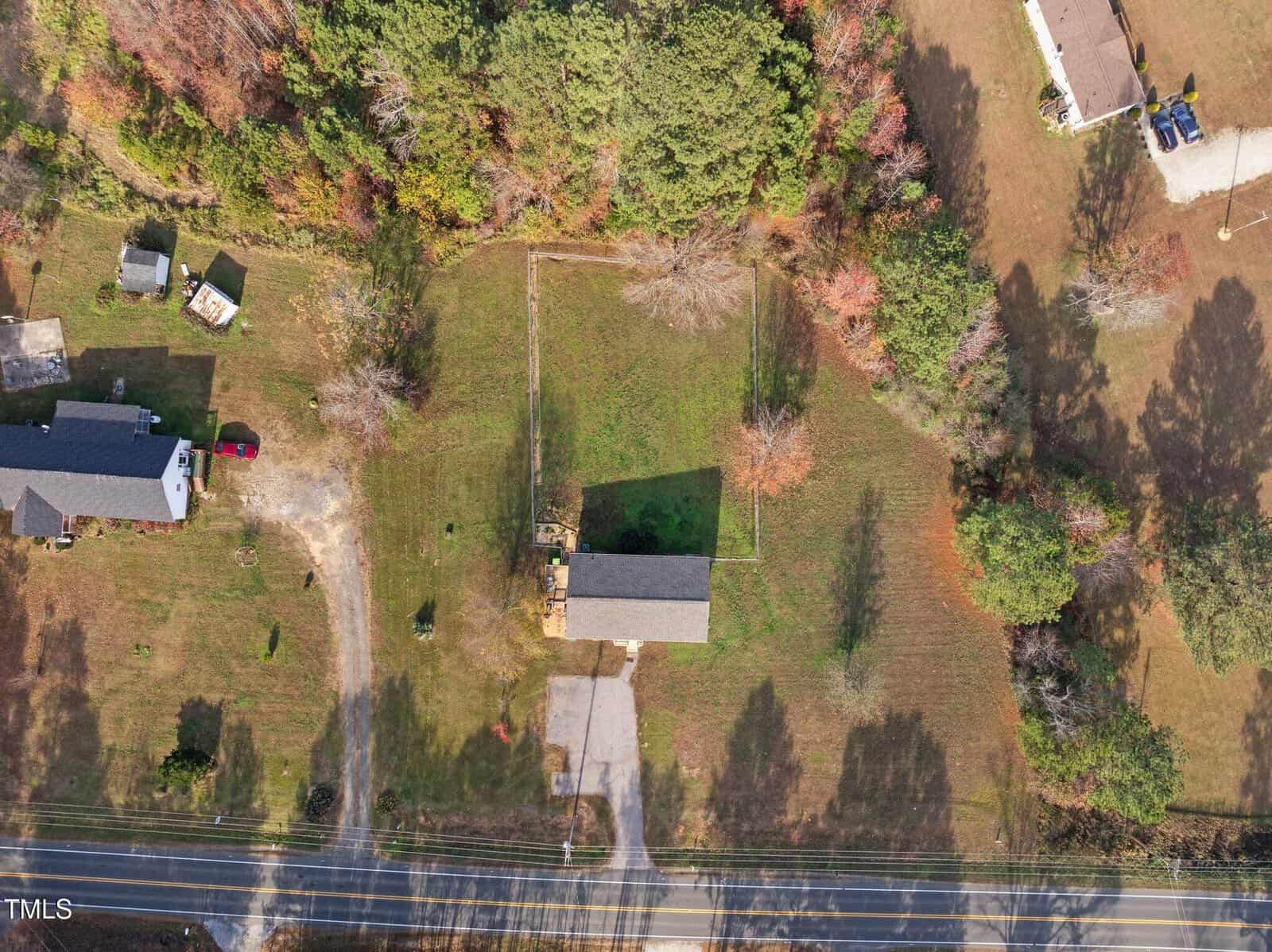 Aerial view of a rural scene with a single-story building surrounded by grass and trees. A driveway leads to the building, and adjacent properties are visible. A road runs horizontally at the bottom of the image.