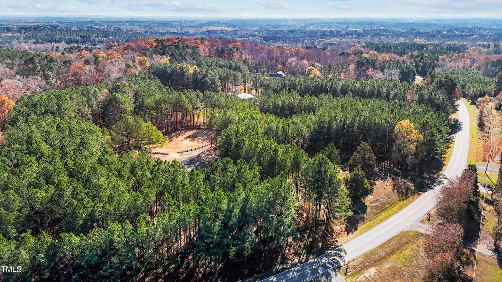 Aerial view of a winding road curving through a dense forest. The trees are a mix of green and autumn colors. A small building is visible in the clearing. The landscape extends towards distant hills under a partly cloudy sky.