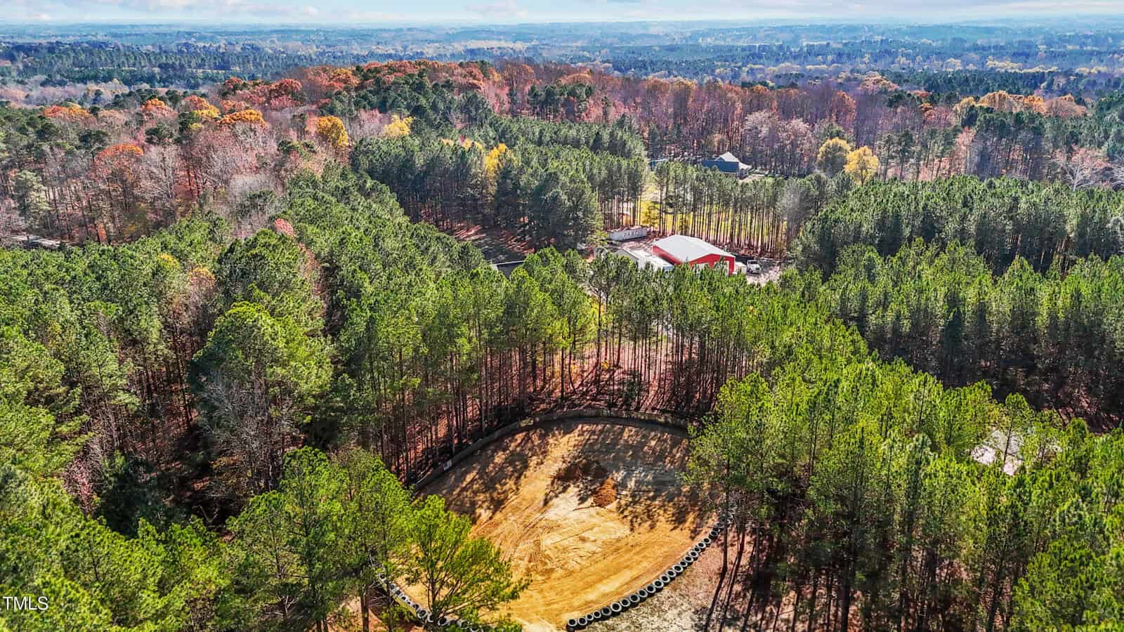 Aerial view of a forested area in autumn with a central dirt track or arena, surrounded by tall trees. A large red-roofed building is visible in the background. The sky is partly cloudy with sunlight filtering through.