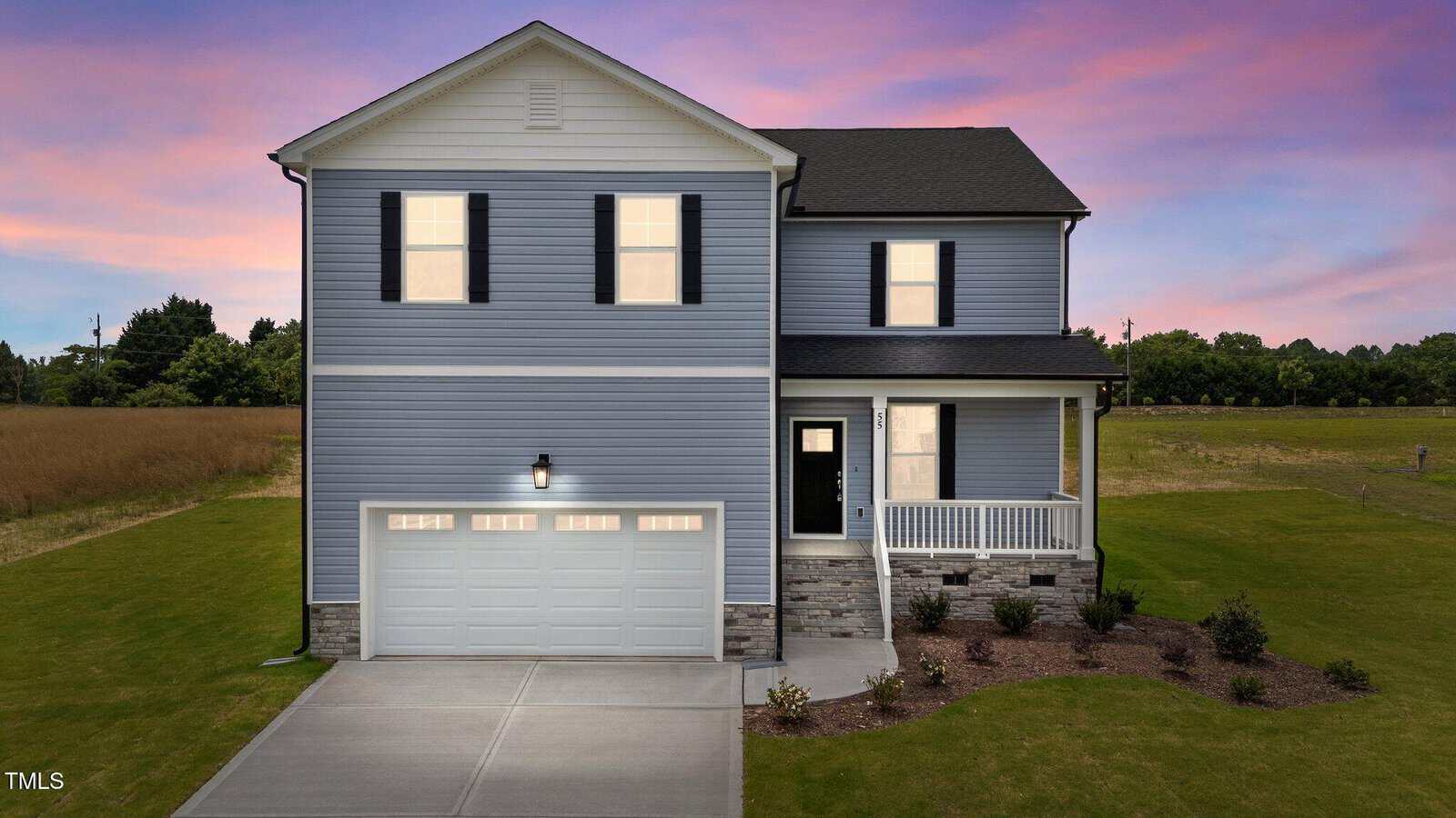 Two-story blue house with black shutters, a covered porch, and a two-car garage. The yard is landscaped with shrubs, and the sky is pink and purple at sunset. The house sits next to an open field.