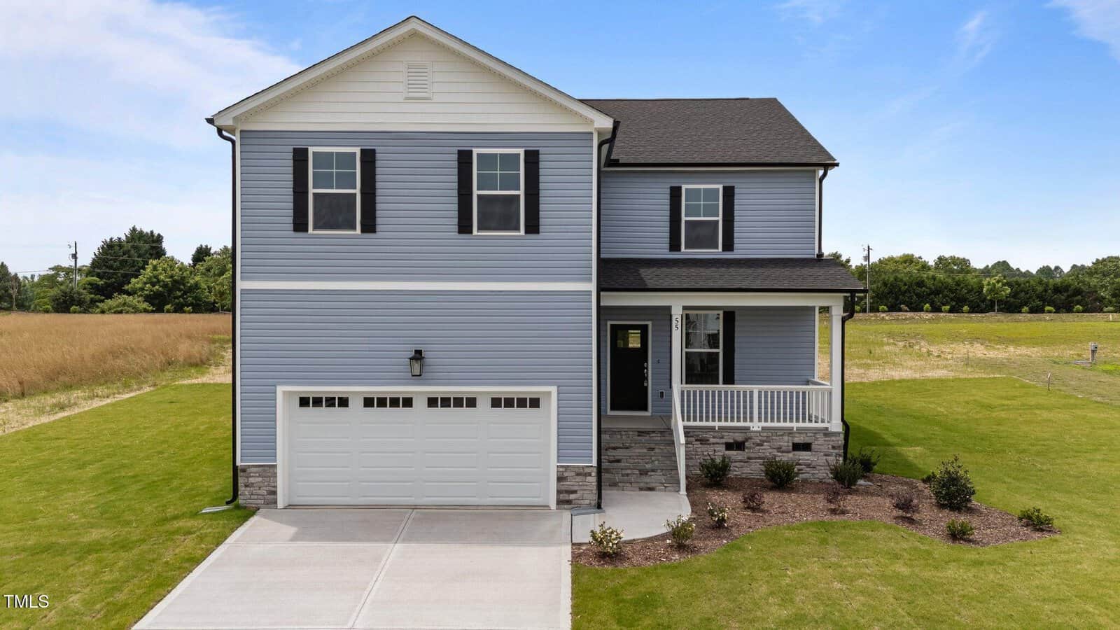 Two-story light blue house with black shutters, a two-car garage, a small covered front porch, and a landscaped yard, set next to an open grassy field under a clear sky.