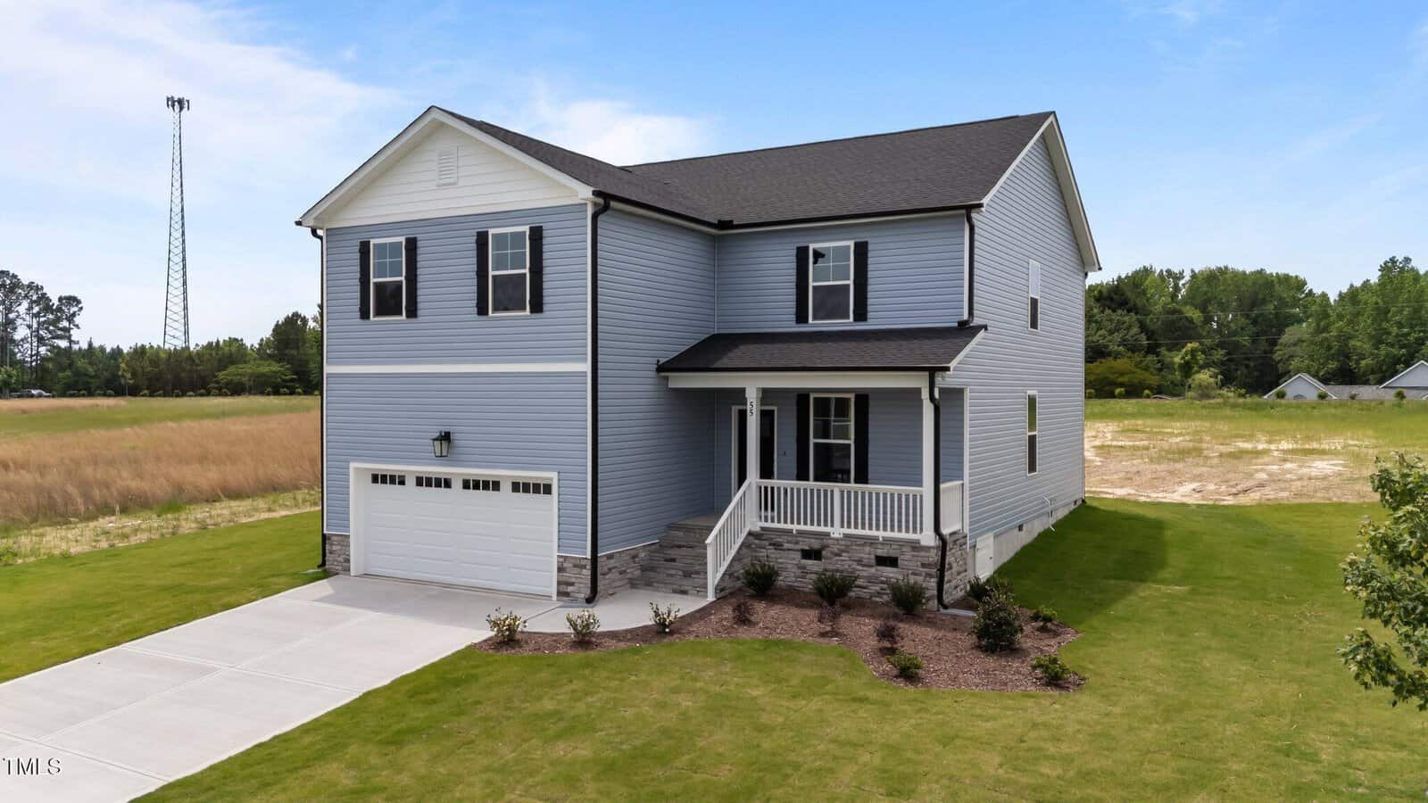 A two-story light gray house with white trim, a covered front porch, and an attached two-car garage. The house sits on a grassy lot with minimal landscaping and a concrete driveway. Trees and an open field are in the background.