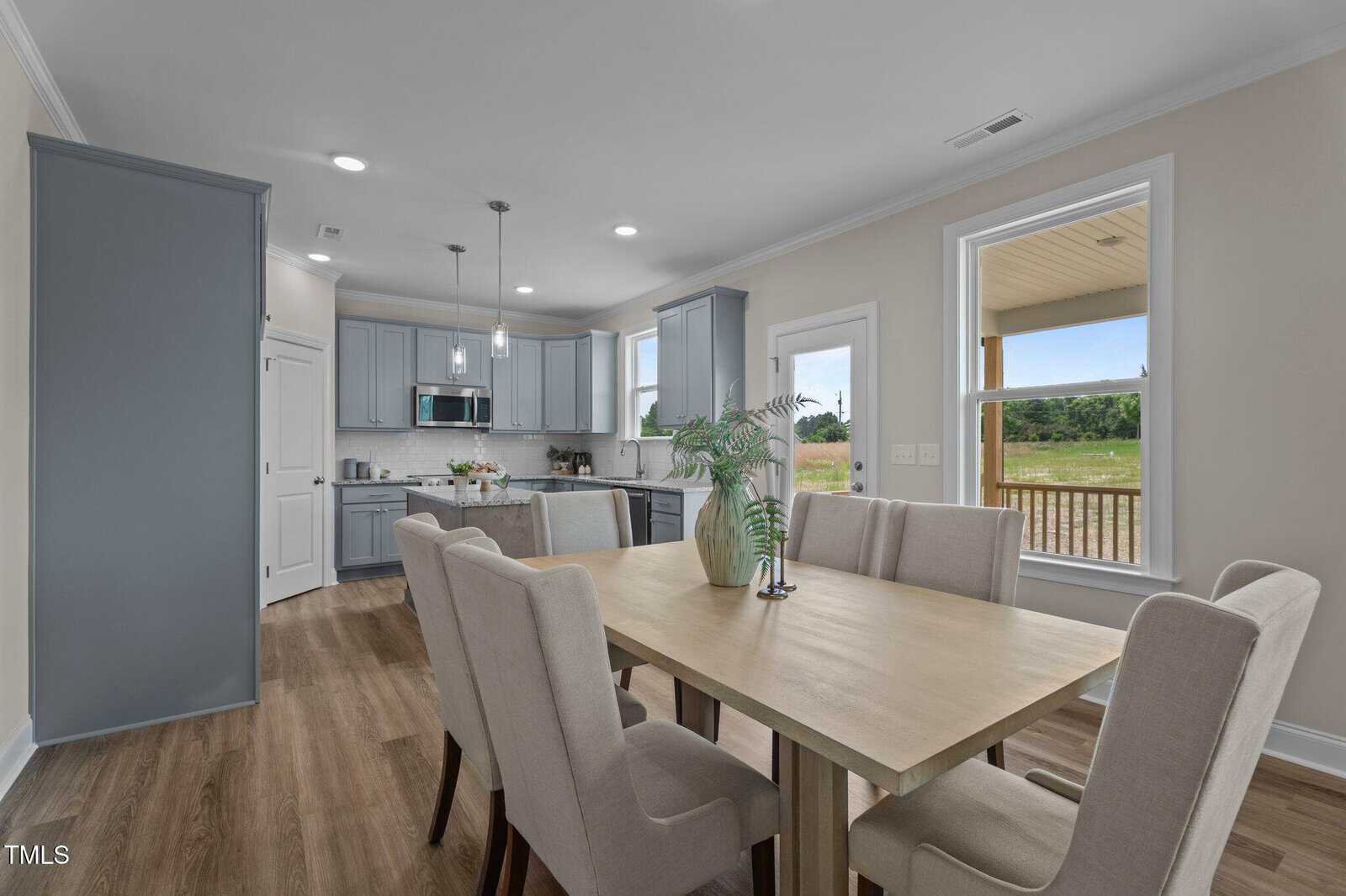 Modern kitchen and dining area with light wood table, six beige upholstered chairs, gray cabinets, stainless steel appliances, and large windows showing a grassy outdoor view. A plant decorates the center of the table.