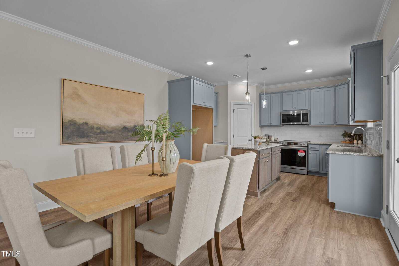 Modern dining area with a wooden table, six beige upholstered chairs, and a vase with greenery. Behind is a kitchen with gray cabinets, stainless steel appliances, and light wood flooring throughout.