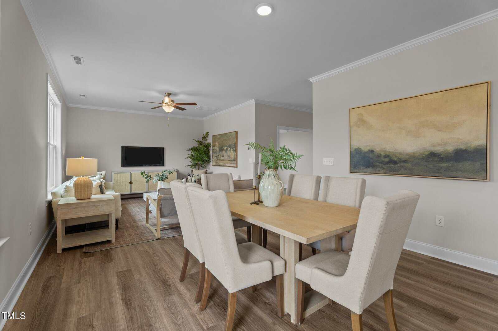 Open-concept living and dining area with neutral walls, light wood flooring, beige upholstered chairs around a wooden table, and a cozy living space with a sofa and TV. Large painting and plant add decorative touches.