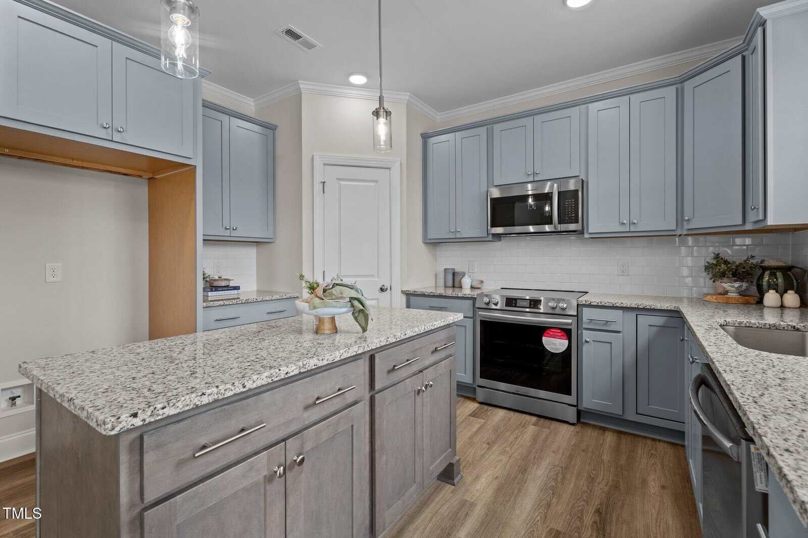 A modern kitchen with light blue cabinets, granite countertops, stainless steel appliances, a built-in microwave, a center island, and wood flooring. There are decorative plants and jars on the counters.