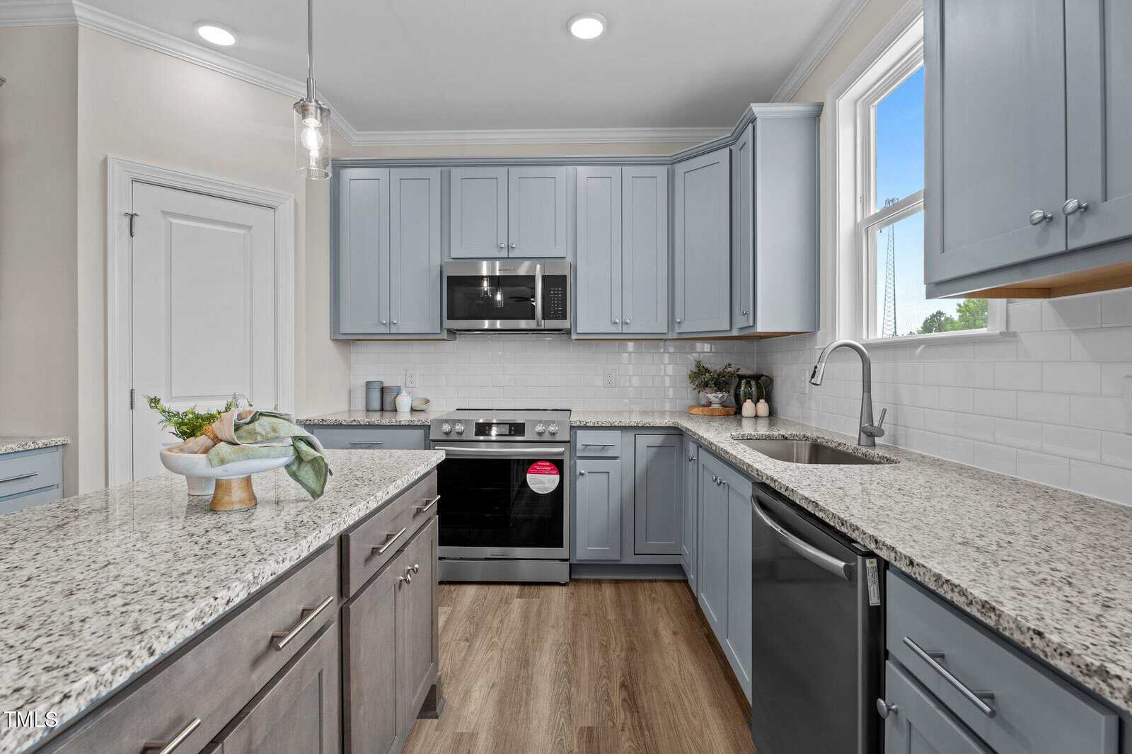Modern kitchen with light gray cabinets, granite countertops, stainless steel appliances, white subway tile backsplash, and wood-look flooring. A window is above the sink and there is a decorative bowl on the island.