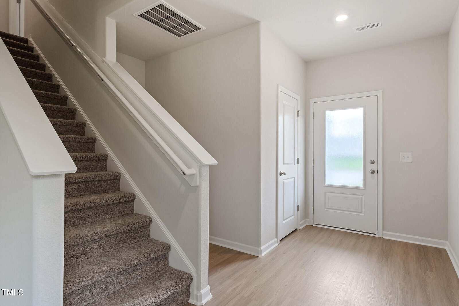Entryway of a house featuring a wooden floor, carpeted stairs to the left with a white railing, a white door with frosted glass panels, and a closed white interior door. Walls are painted light beige, and there's a white air vent above.