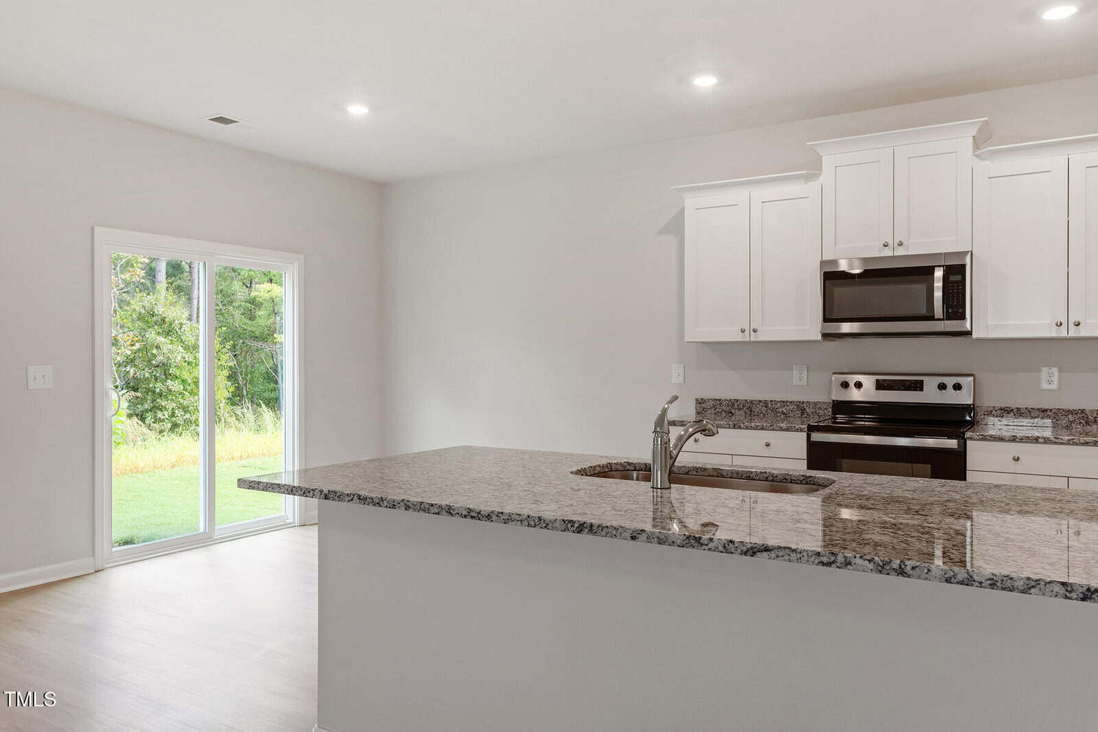 A modern kitchen with white cabinets and stainless steel appliances, including an oven and microwave. A granite countertop island features a built-in sink. Sliding glass doors on the left open to a view of greenery outside. The space is well-lit.