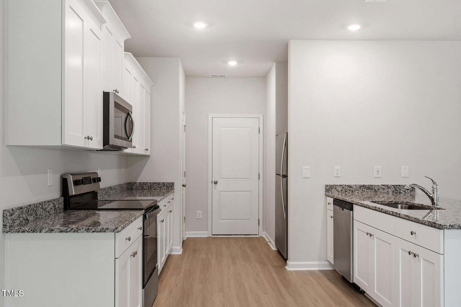 Modern kitchen with white cabinetry and granite countertops. Features stainless steel appliances, including a microwave and stove. Hardwood flooring and a recessed lighting fixture create a clean, minimalist look. A door and sink area are also visible.