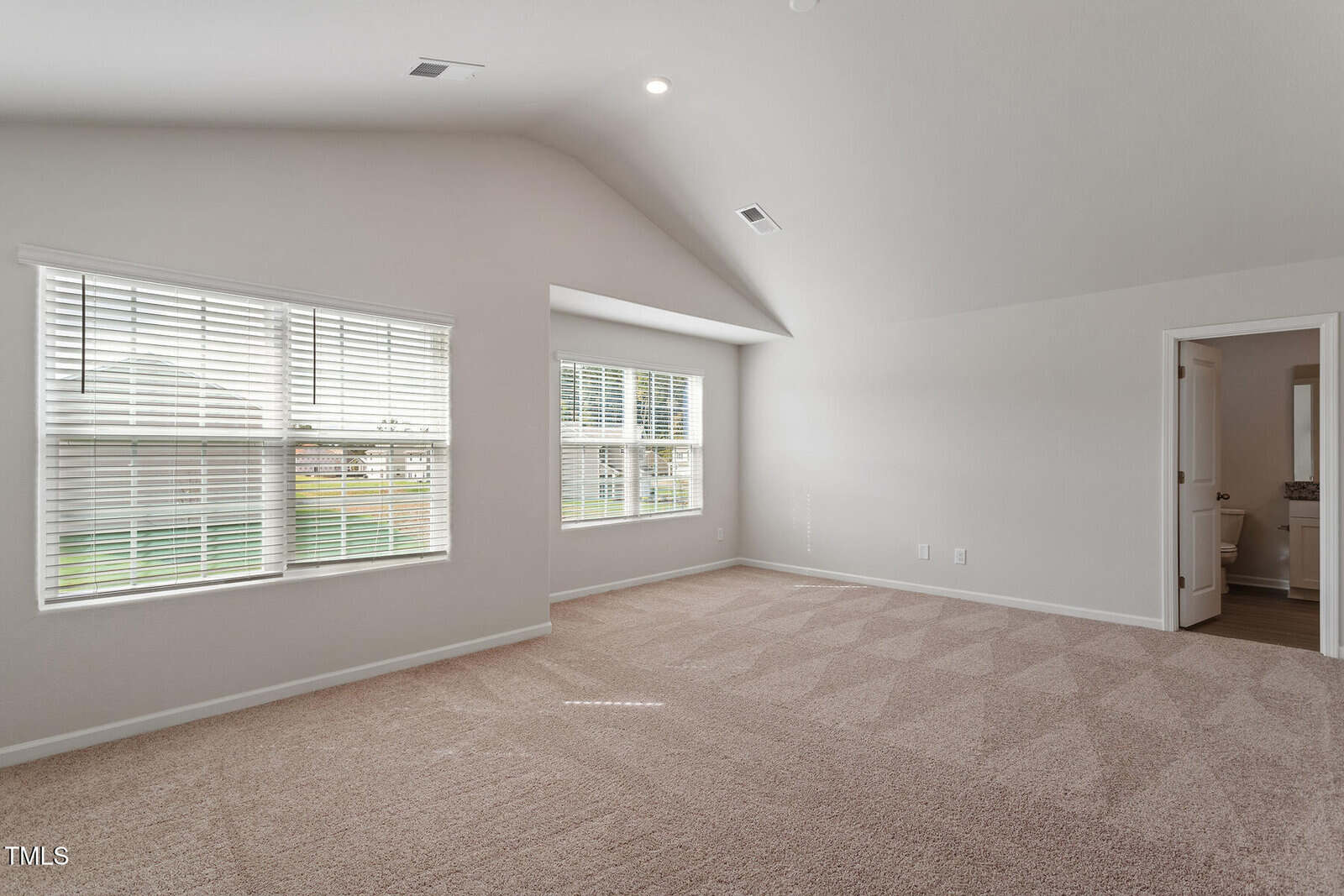 Spacious, empty room with cream-colored walls and carpeted floor. Large windows let in natural light. A door on the right leads to a small bathroom with visible fixtures. The ceiling is vaulted, adding to the open feel.