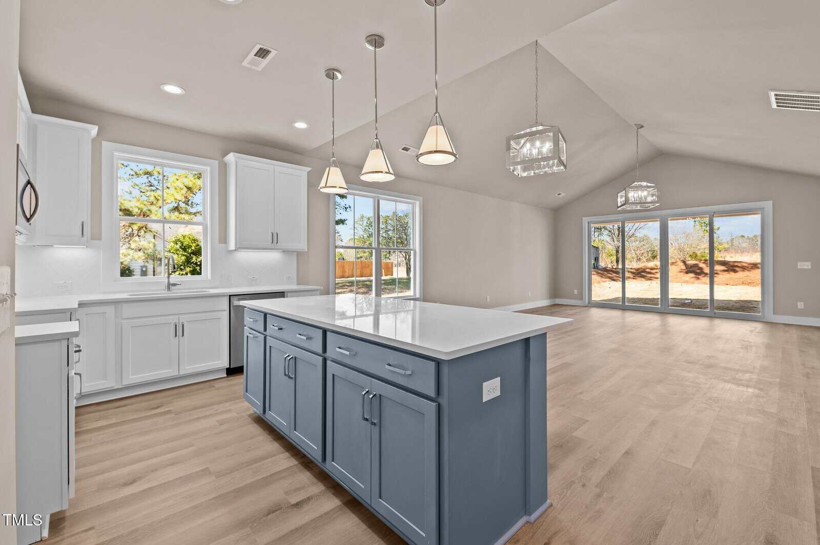 Modern open-concept kitchen and living area with light wood floors, white cabinets, a blue island with a white countertop, pendant lighting, and large windows and sliding doors overlooking a yard.