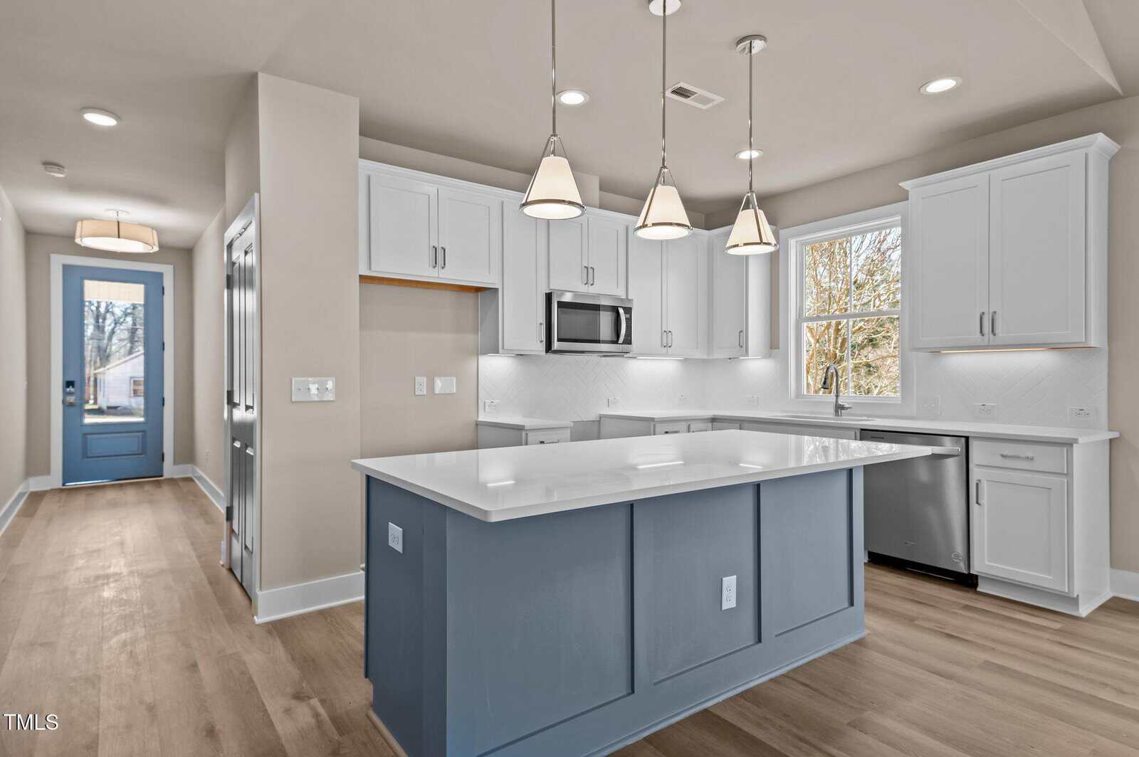 Modern kitchen with white cabinets, gray-blue island, pendant lights, stainless steel appliances, wood flooring, and a window letting in natural light. A blue door and hallway are visible in the background.