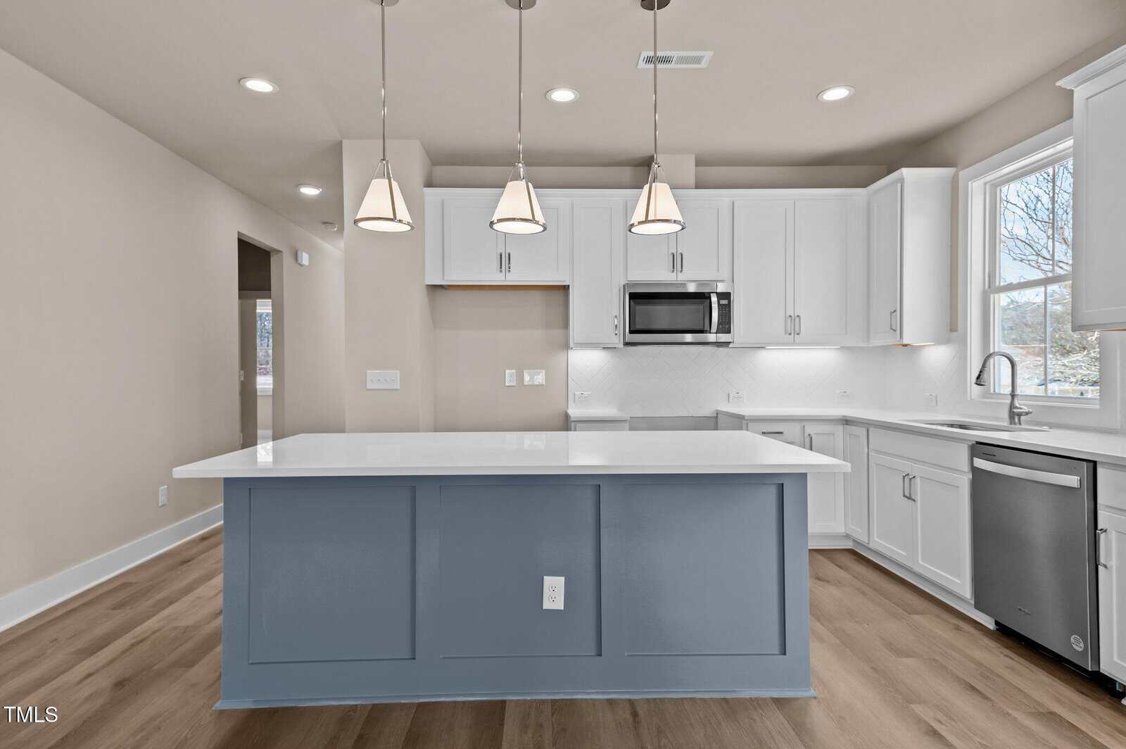 A modern kitchen with white cabinets, stainless steel appliances, a large blue island with a white countertop, pendant lighting, light wood flooring, and a window above the sink.
