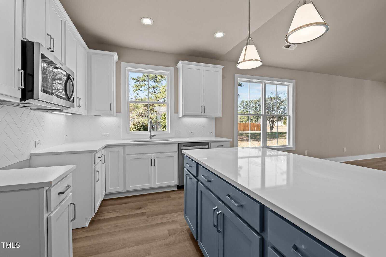 Modern kitchen with white cabinets, blue island, quartz countertops, stainless steel appliances, pendant lights, large windows, and light wood flooring. Bright, open space with outdoor view.