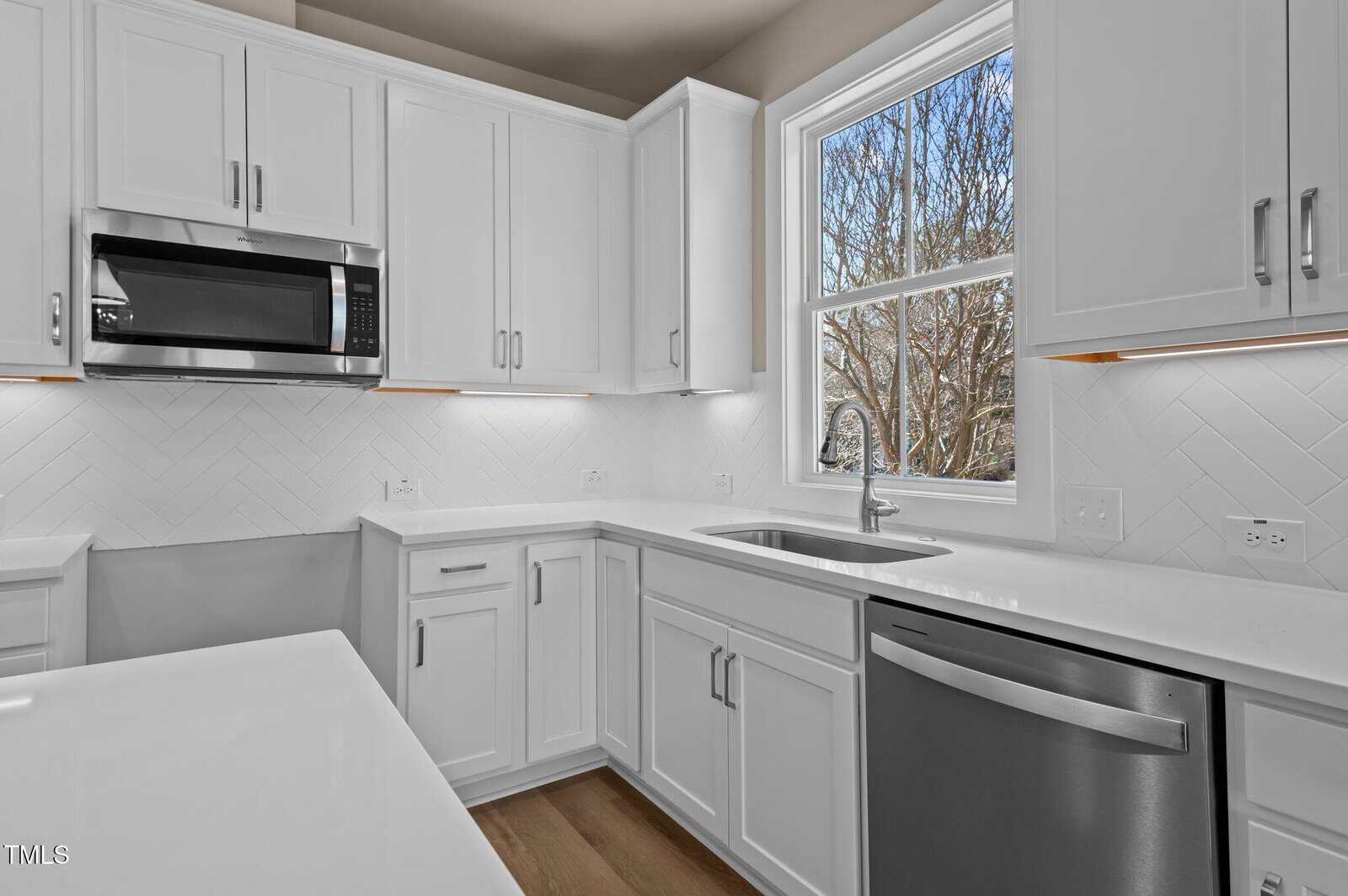 Modern kitchen with white cabinets, stainless steel appliances, a built-in microwave, and a dishwasher. There is a window above the sink with a view of tree branches outside. The countertops are white with a clean finish.