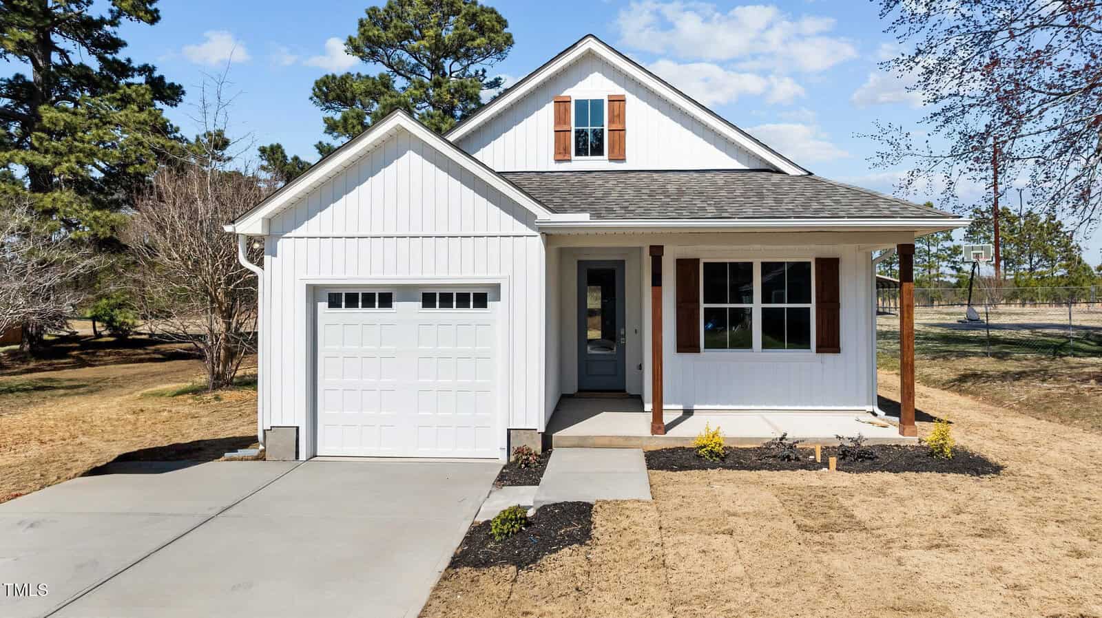 A small white house with a front porch, single-car garage, pitched roof, and brown accents. The yard has sparse grass and some young plants, with a concrete driveway and sidewalk leading to the entrance.