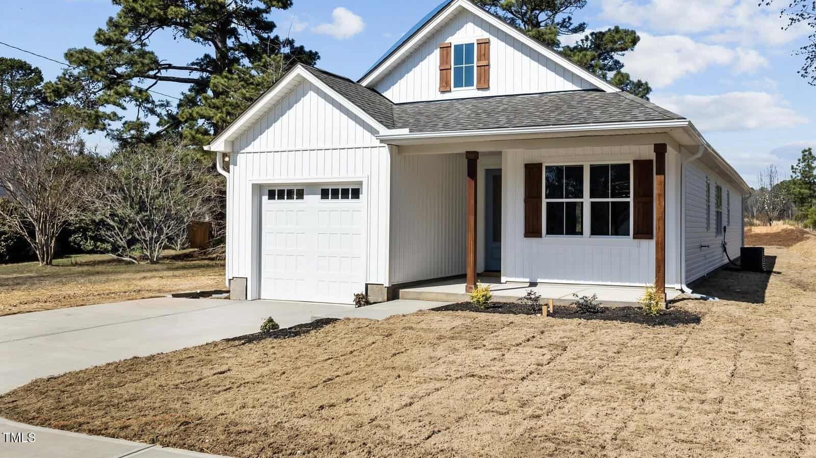 A newly constructed white single-story house with a small covered porch, wooden shutters, attached single-car garage, concrete driveway, and freshly laid sod in the front yard. Tall trees and blue sky are in the background.