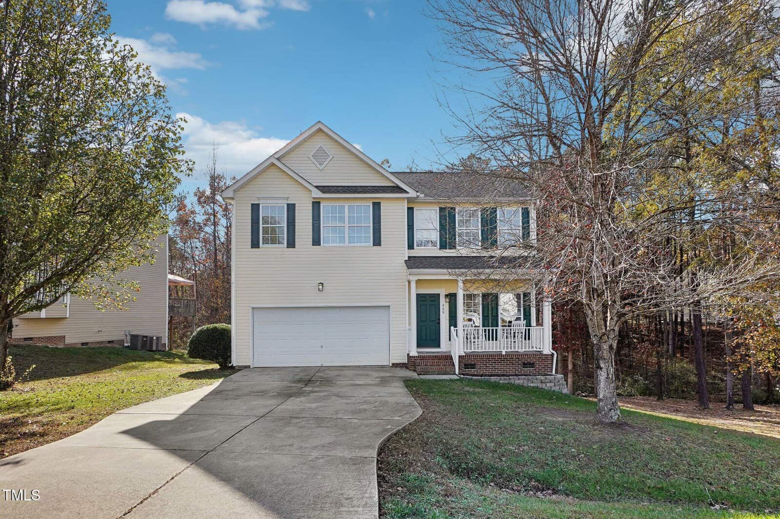 A charming two-story house at 459 Mayview Drive in Creedmoor features beige siding and green shutters. It boasts a two-car garage and a lovely brick front porch. The driveway meanders through an autumn-touched yard, while the sky above is a stunning blue with scattered clouds.