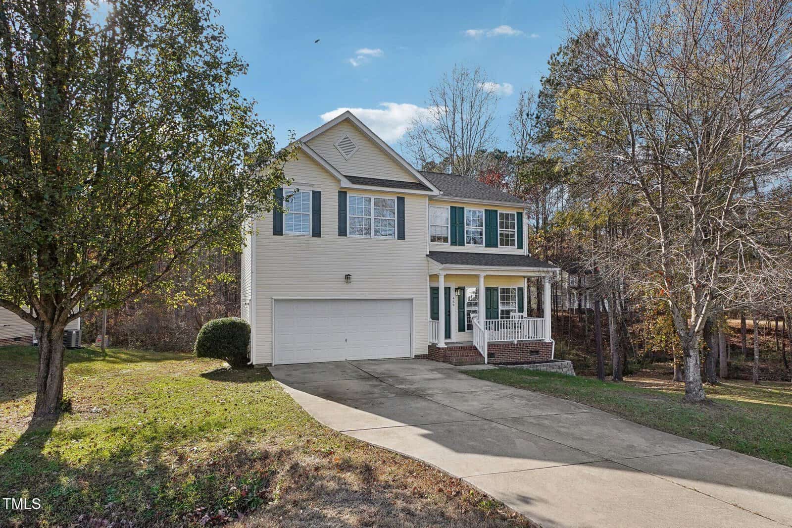A two-story house with beige siding, a double garage, and a small front porch. It is surrounded by trees and has a paved driveway. The sky is clear and the yard is grassy with some fallen leaves.