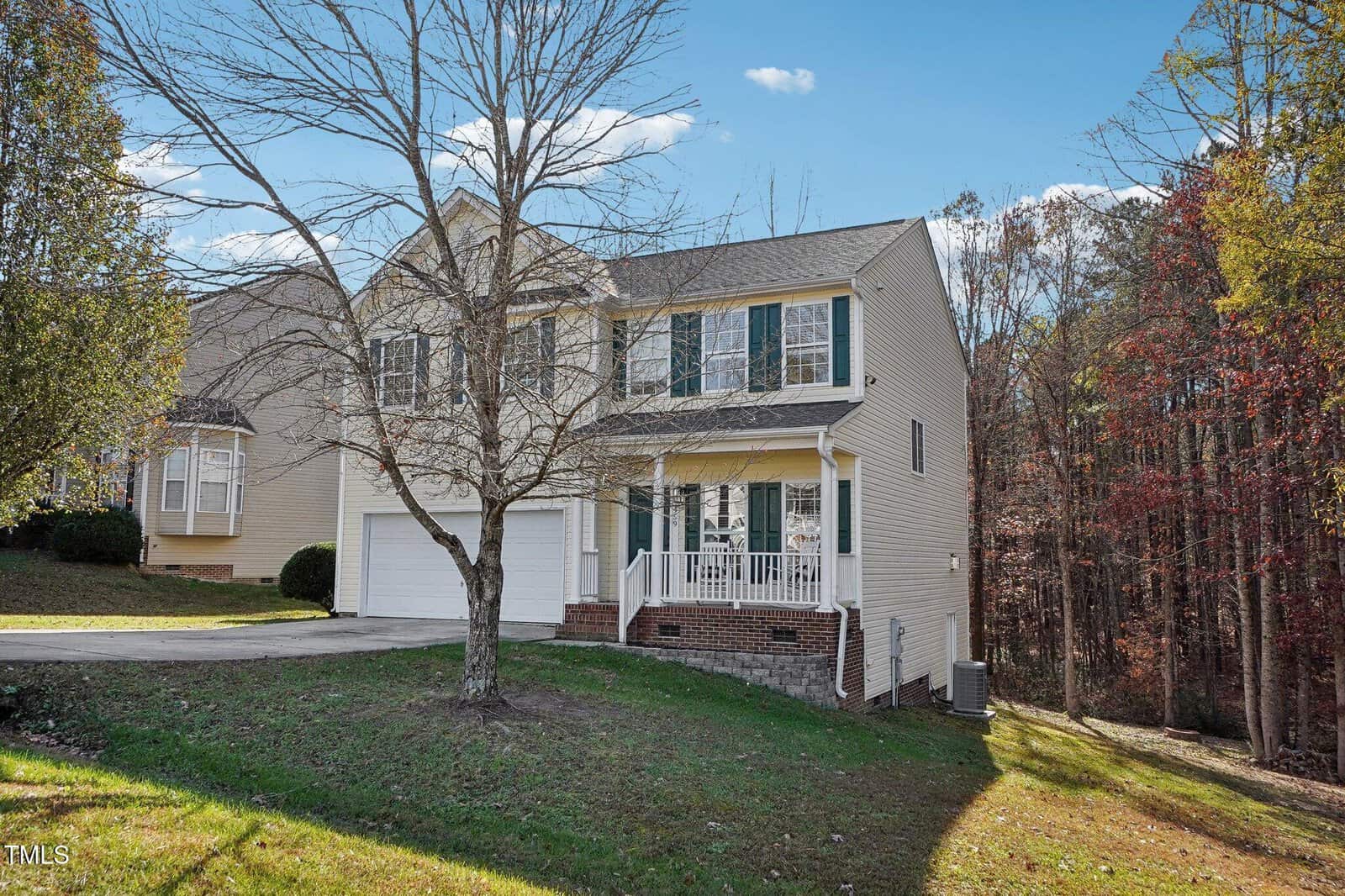 A beige two-story house with a two-car garage and a leafless tree in the front yard. The house has white trim and shutters and sits on a grassy slope with woods in the background. The sky is clear with a few clouds.