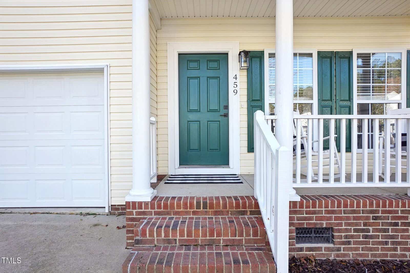A front entrance of a house featuring a green door with the number 459. There is a small porch with white railing and brick steps leading up to it. The house has light-colored siding and a garage door to the left.