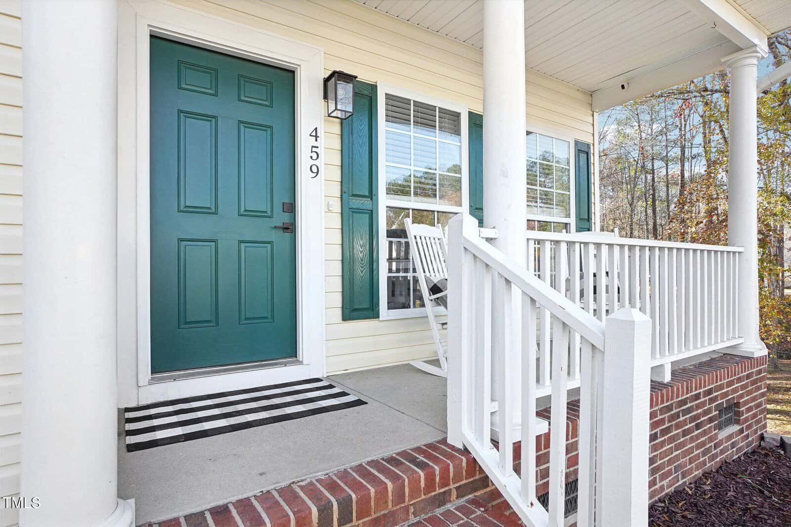 A porch with white railings and pillars, featuring a green door with the number 459. There's a black and white striped doormat and two white rocking chairs. Beige siding and a window with closed blinds are visible in the background.