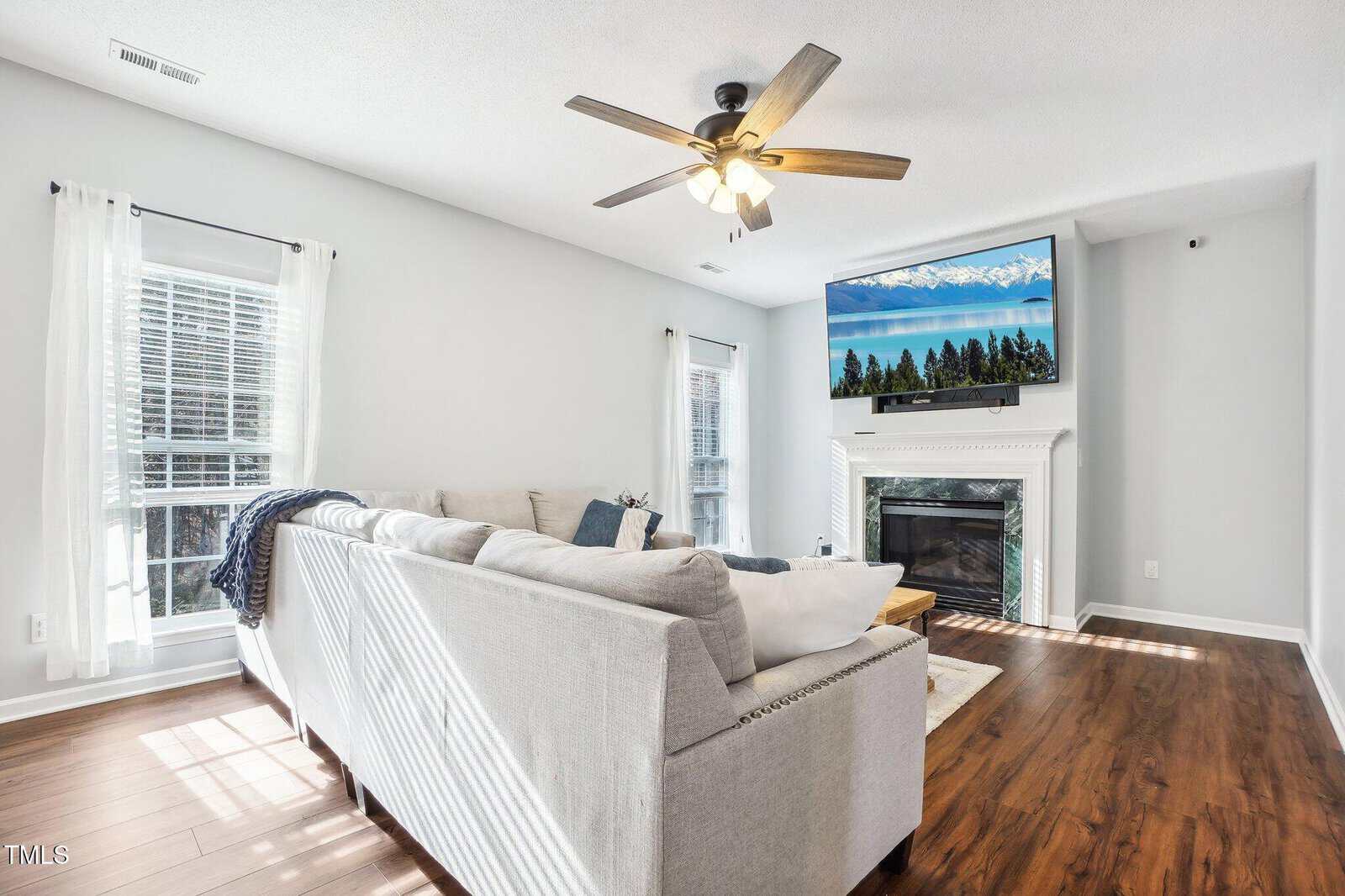 A living room with a light gray sectional sofa, a ceiling fan with lights, and a mounted television above a fireplace. The walls are light-colored, and windows with sheer curtains allow natural light to illuminate the space. The floor is hardwood.
