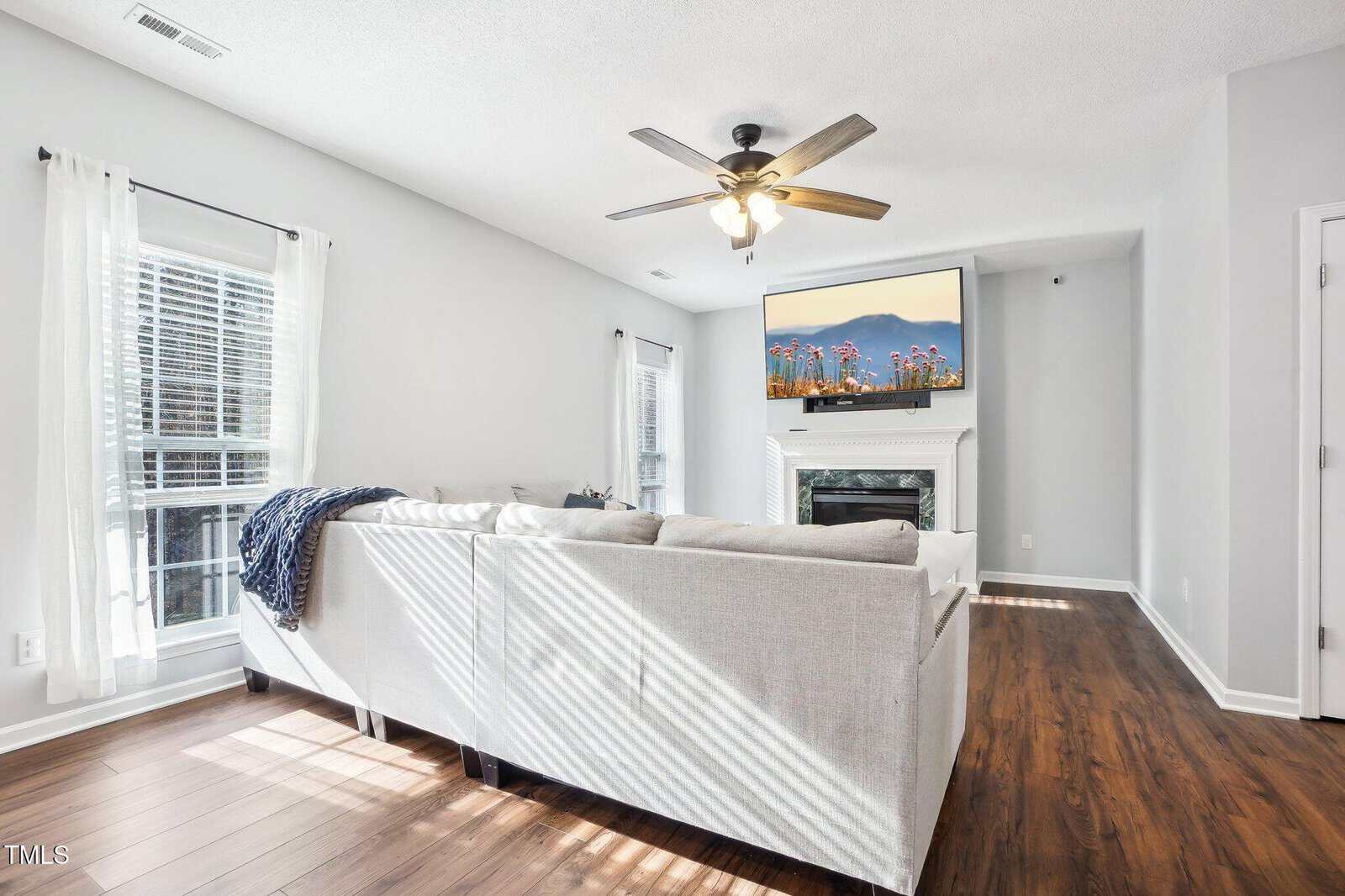 A bright living room with a large gray sectional sofa, a ceiling fan, and a mounted TV above a fireplace. Sunlight streams in through two windows with white curtains. The floor is hardwood, and the walls are painted light gray.