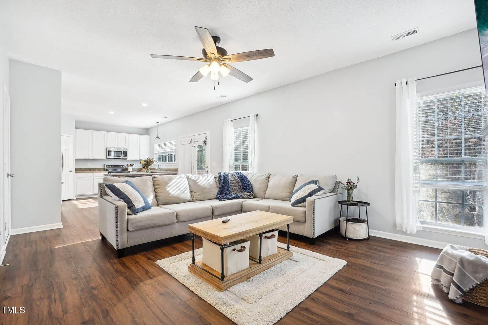 A spacious living room with a large beige sectional sofa, wooden coffee table, and a ceiling fan. The room has wood flooring and is adjacent to a kitchen area. Natural light comes through two tall windows with white curtains.