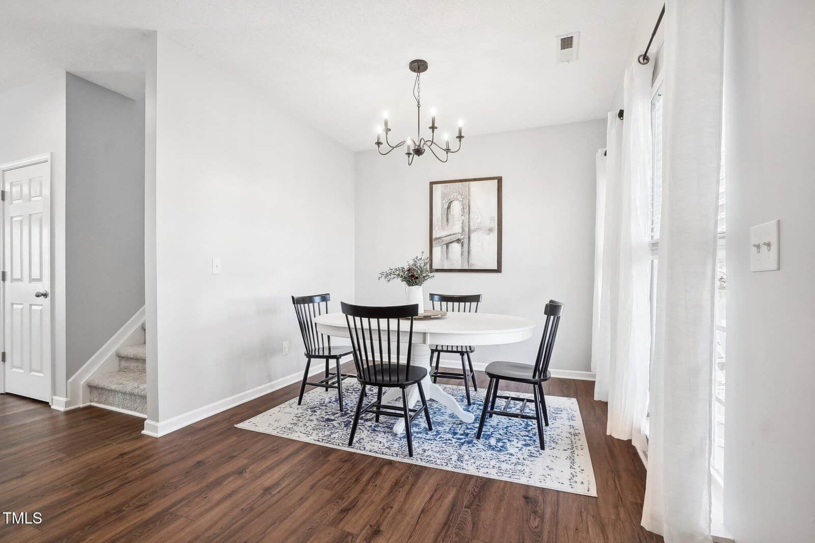 A dining room with a round white table, five black chairs, and a blue-patterned rug. A chandelier hangs above the table, and a framed artwork is on the wall. Curtains cover windows on the right, and a staircase is visible on the left.
