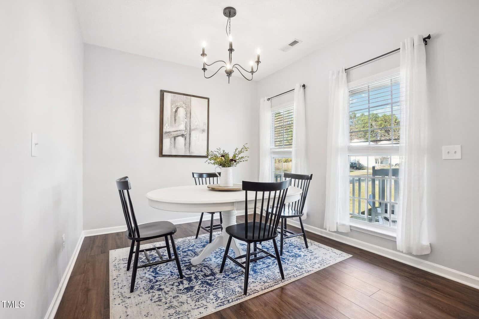 A dining room with a round white table and four black chairs on a blue patterned rug. A chandelier hangs above the table. A large framed picture is on a light gray wall. Two windows with white curtains allow natural light into the room.