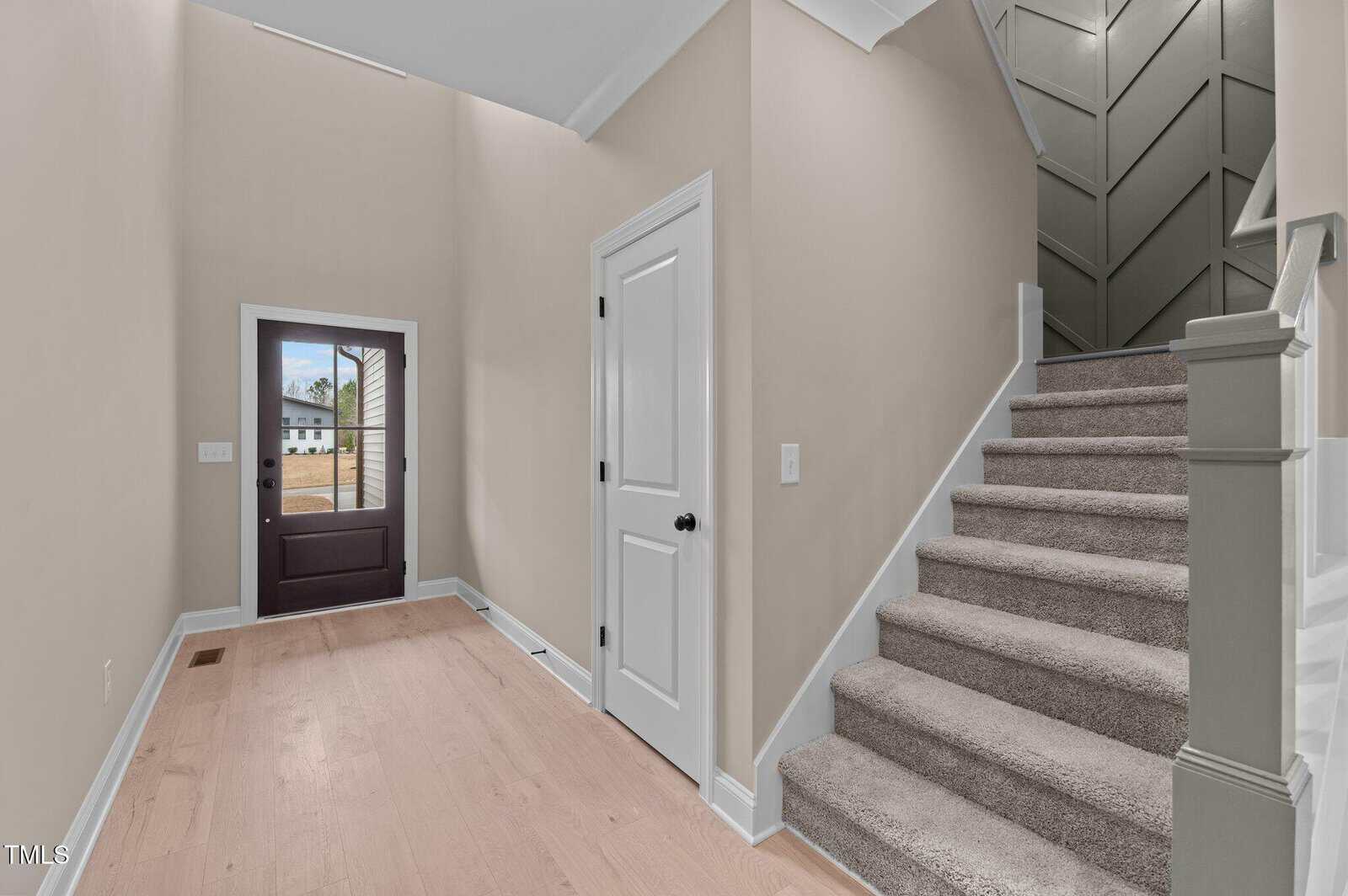 A bright entryway with light wood flooring, beige walls, a dark front door with glass panels, a closed white door, and carpeted stairs leading up next to a green accent wall with a geometric pattern.