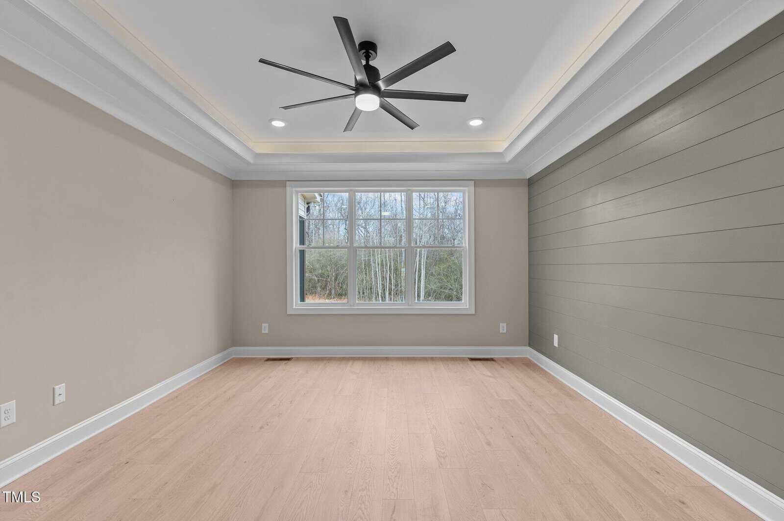 Empty room with light wood flooring, beige and green walls, a large window showing trees outside, a tray ceiling with recessed lighting, and a modern black ceiling fan.