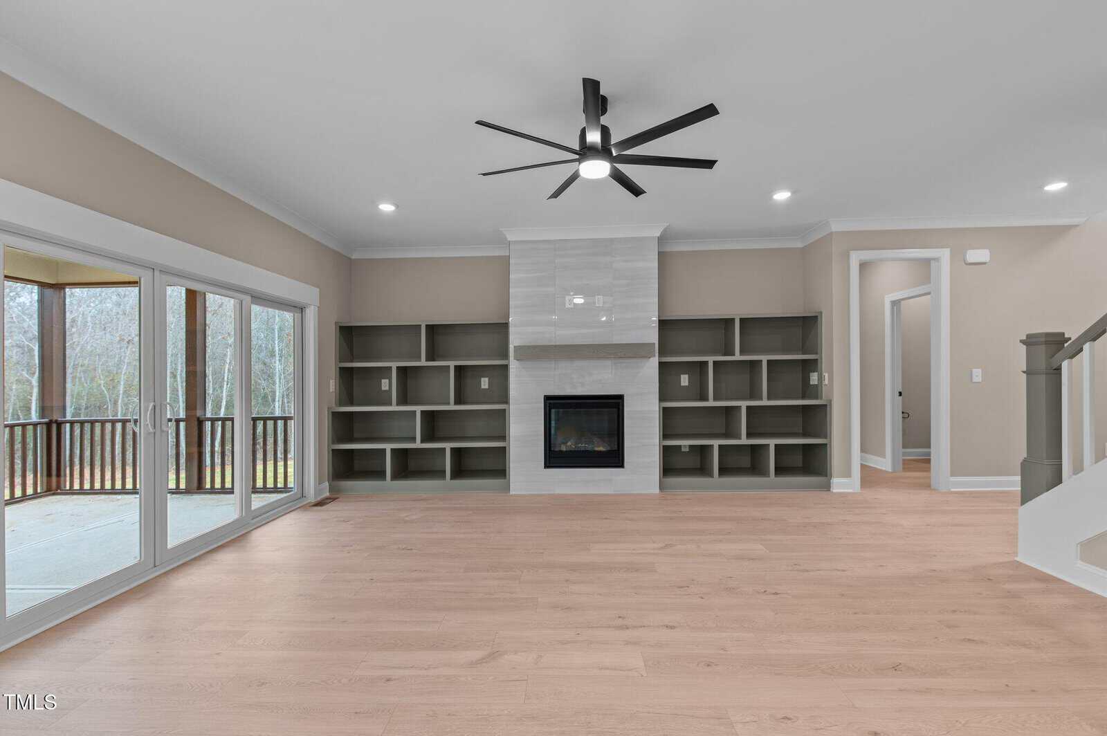 A modern living room with light wood floors, built-in shelves on both sides of a central fireplace, a ceiling fan above, large sliding glass doors to a covered patio, and a staircase to the right.