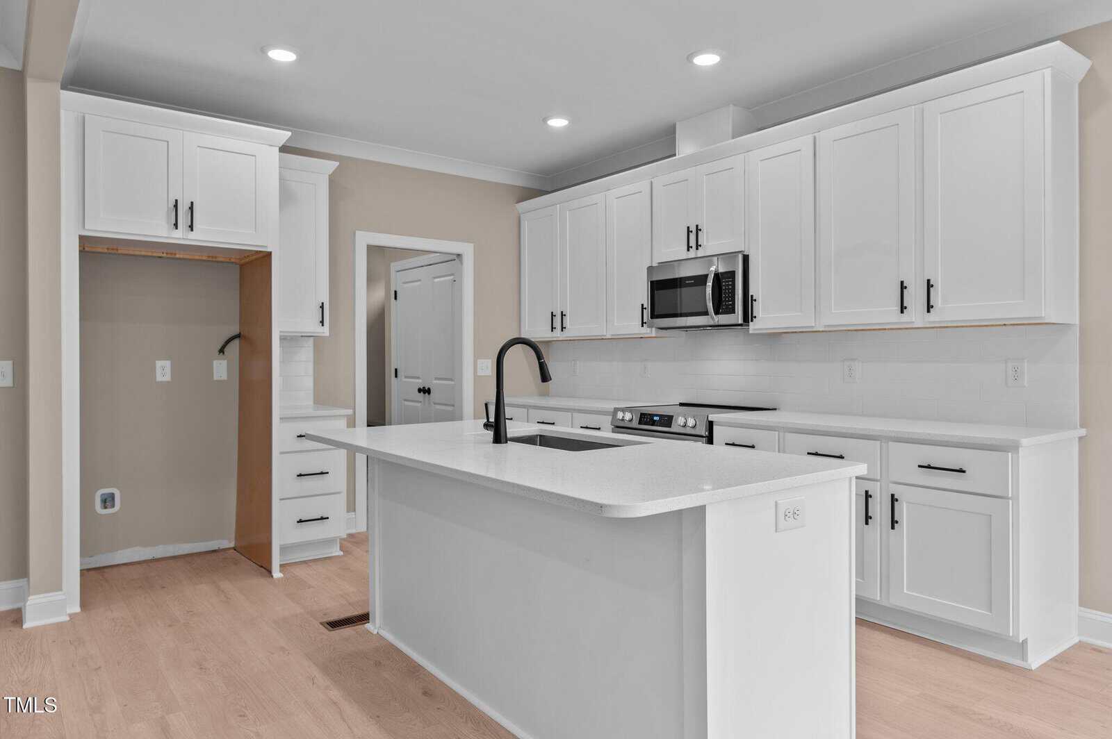 Modern kitchen with white cabinets, black hardware, a large island with a built-in sink and faucet, light wood floors, stainless steel microwave, and recessed lighting. An empty alcove is visible for a refrigerator.