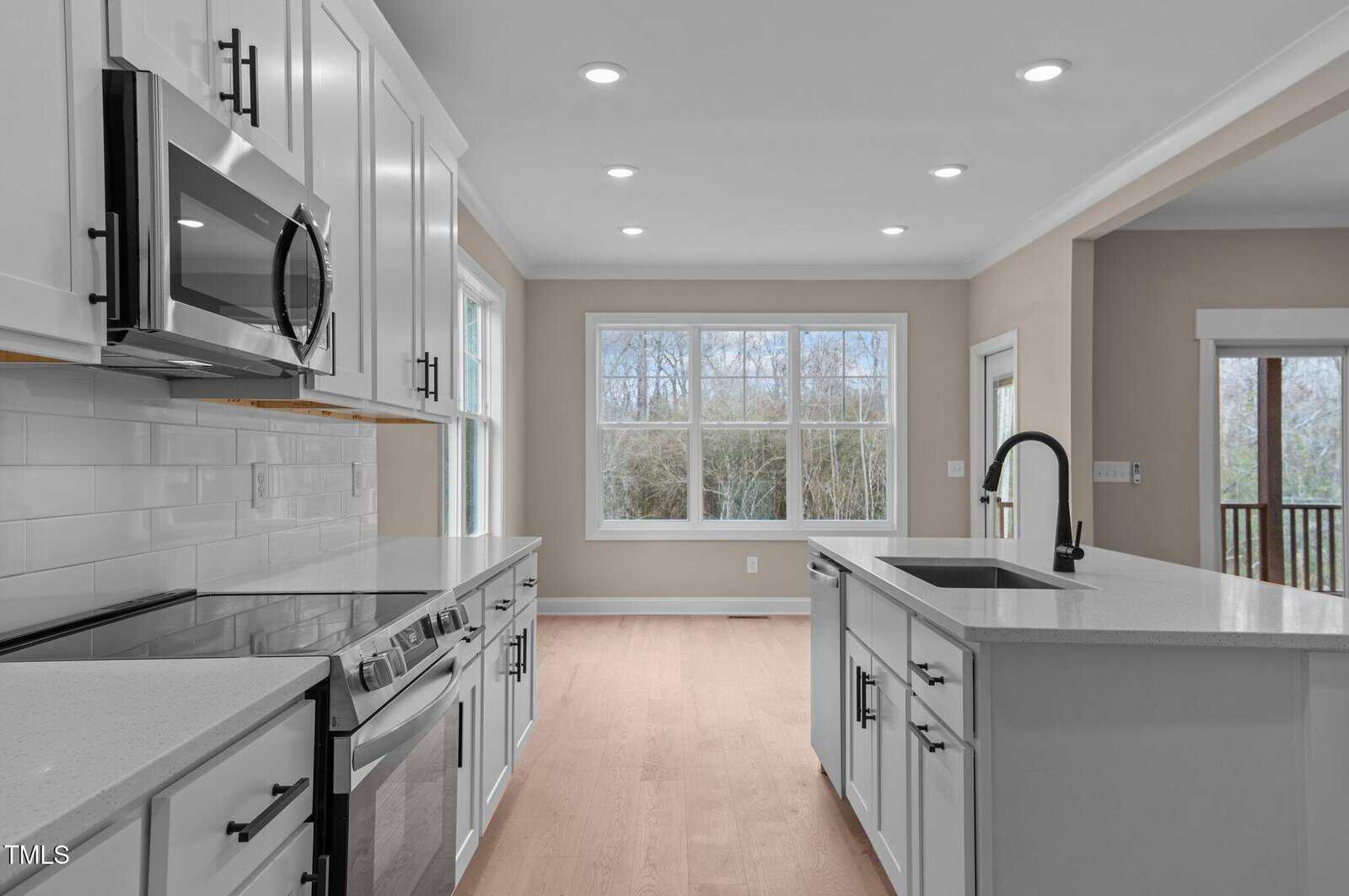 Modern kitchen with white cabinets, stainless steel appliances, subway tile backsplash, and an island with a black faucet. Bright dining area with large windows offering views of trees outside. Light wood flooring throughout.