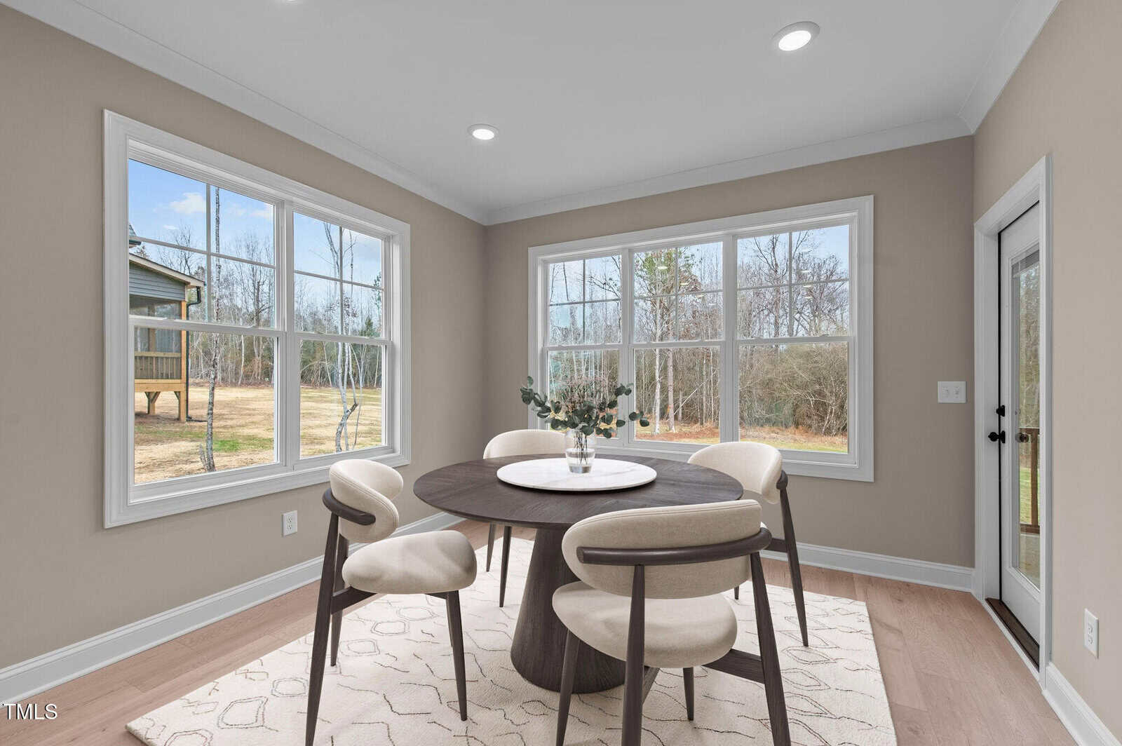 A bright dining area with large windows, beige walls, a round wooden table, four cushioned chairs, a white rug, and a vase with greenery. The windows reveal a view of the outdoors with trees and dry grass.