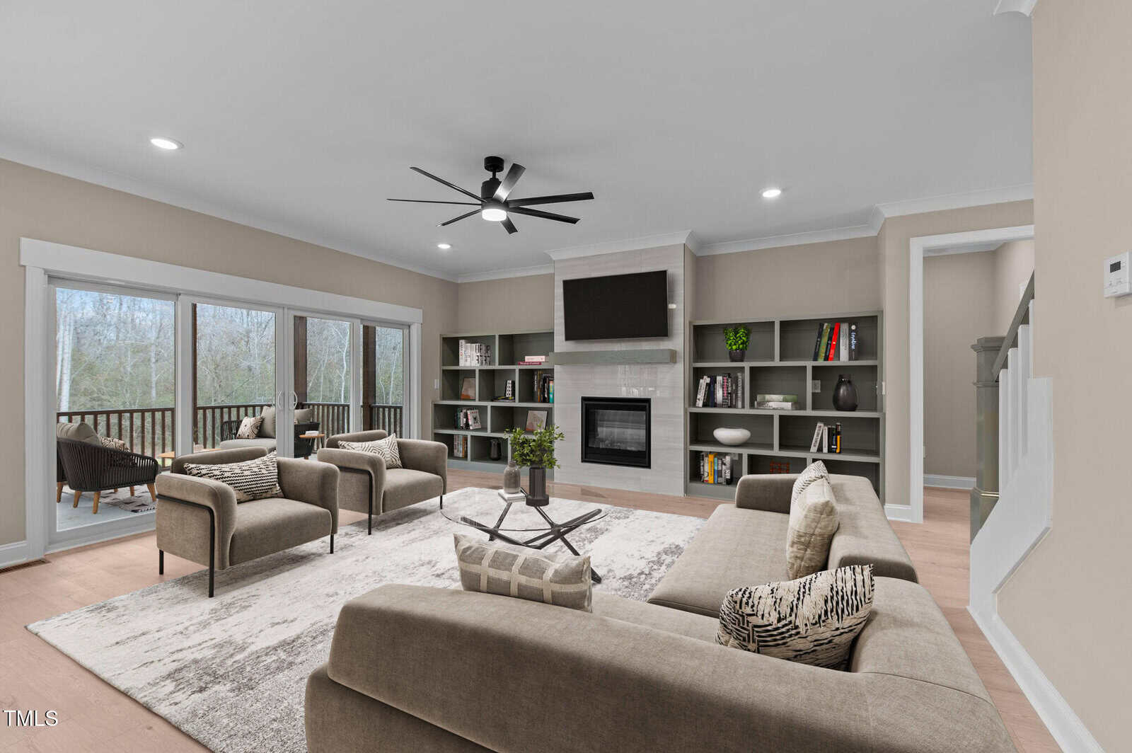 A modern living room with beige walls, a gray sectional sofa, two armchairs, a coffee table, built-in shelves, a fireplace with a TV above it, large windows, and a view of a wooded deck outside.