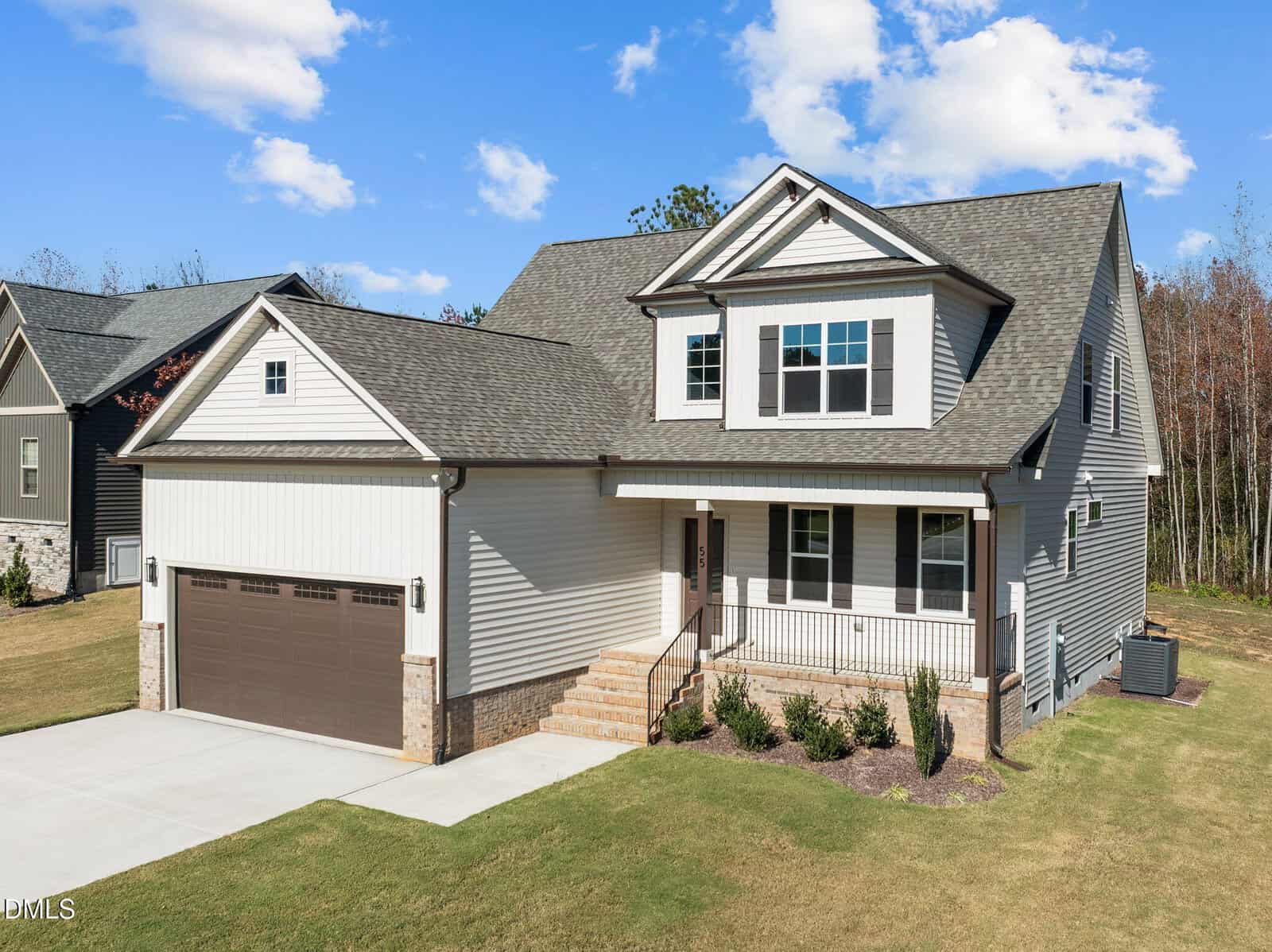 A modern two-story house with gray siding, a brown double garage door, covered front porch, concrete driveway, and neatly landscaped yard, set against a backdrop of trees and a blue sky with scattered clouds.
