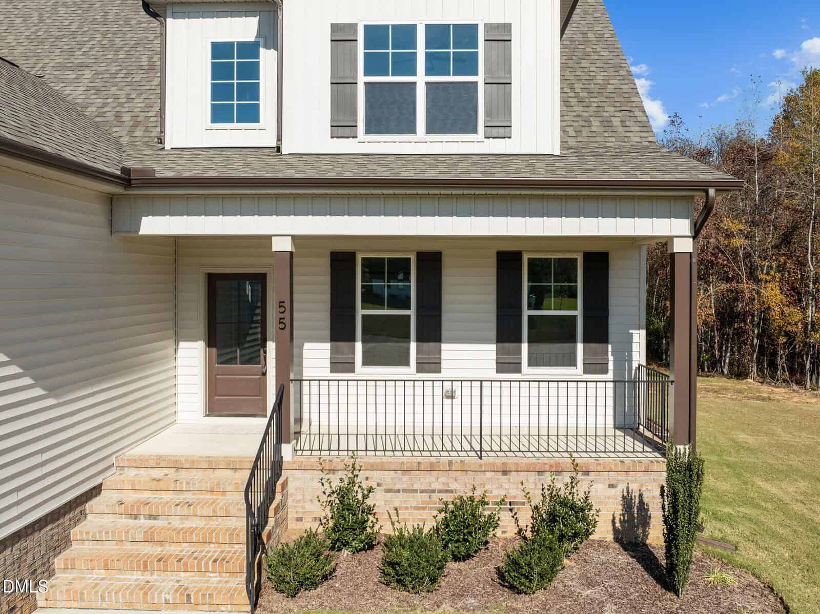 A modern house with light-colored siding, a front porch with black railings, steps leading to a brown door, three front windows with shutters, and a small landscaped area with shrubs.