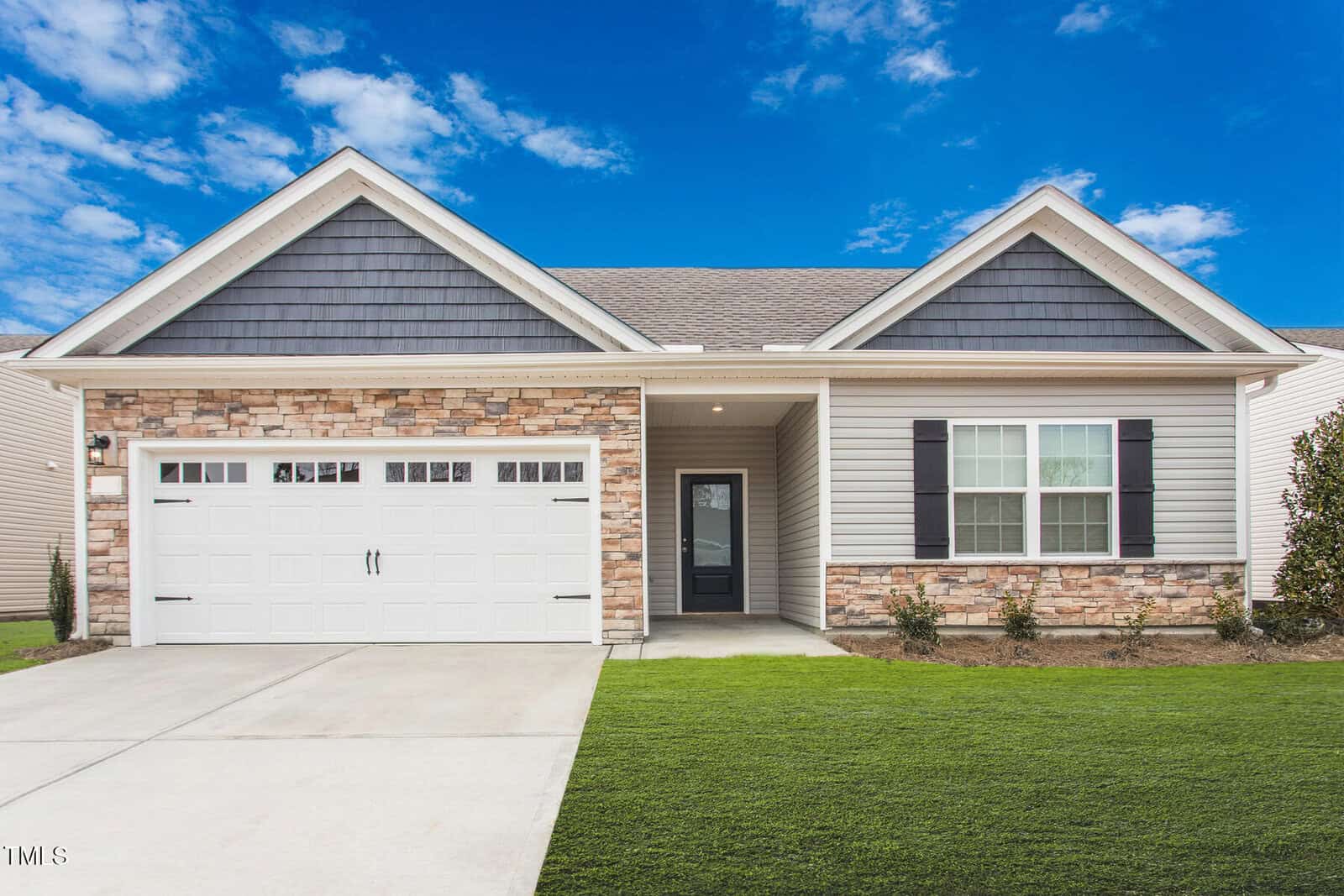 A single-story suburban house with a two-car garage, stone and siding exterior, dark roof shingles, white trim, and a manicured lawn under a bright blue sky with scattered clouds.