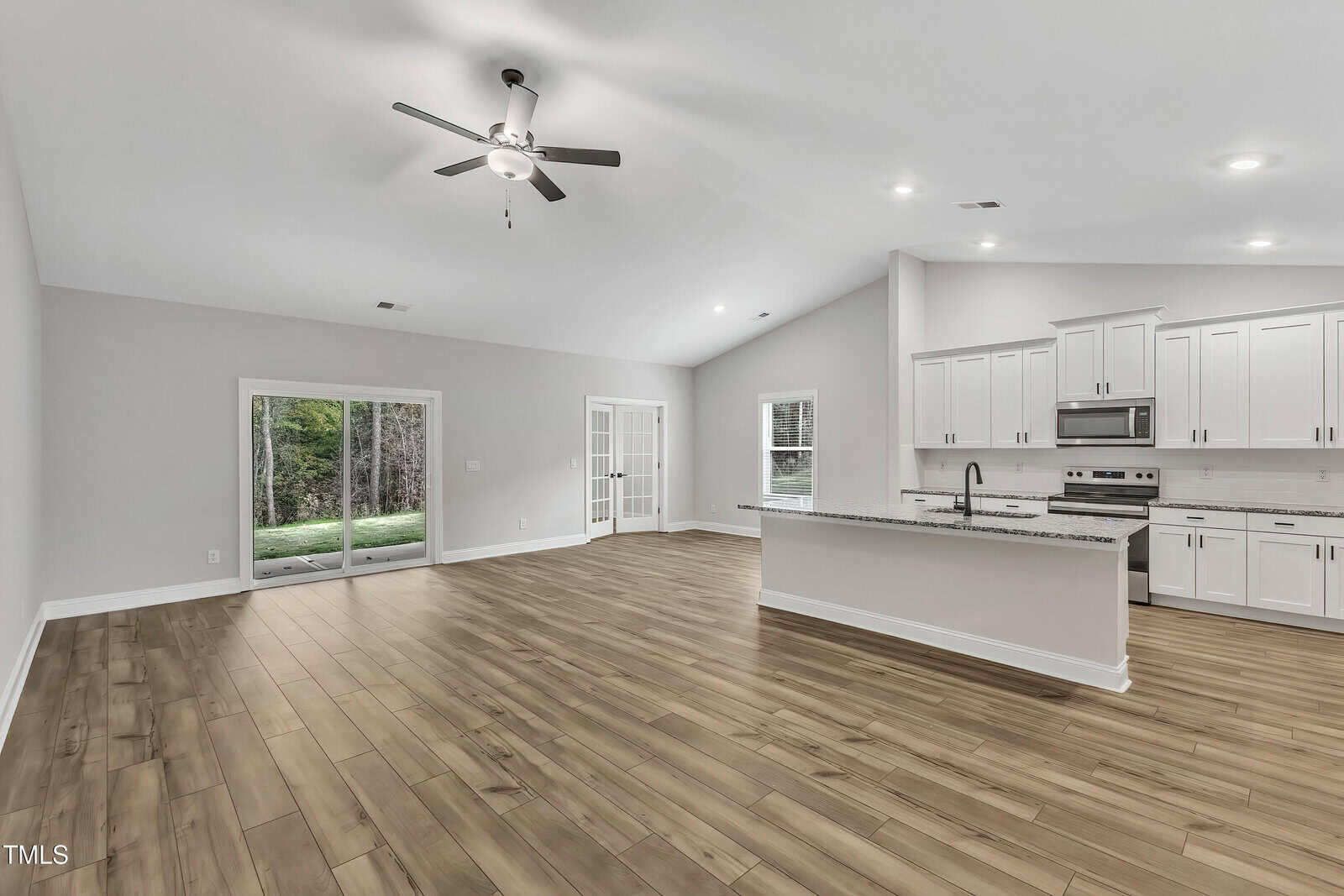 Spacious open-concept kitchen and living area with light wood flooring, white cabinets, stainless steel appliances, recessed lighting, ceiling fan, sliding glass doors, and a large window.