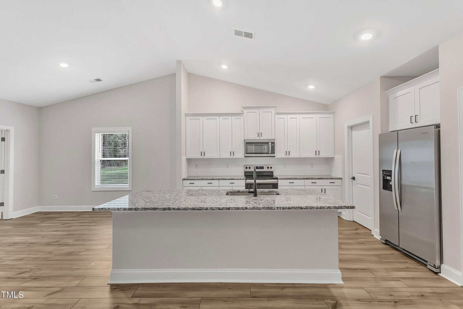 Modern kitchen with white cabinets, stainless steel appliances, a large center island with a granite countertop, and wood-style flooring. The room is well-lit with recessed lighting and natural light from a window.