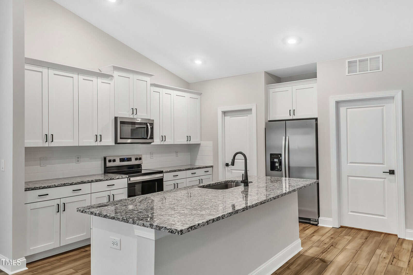 A modern kitchen with white cabinets, stainless steel appliances, a built-in microwave, an electric stove, a double-door refrigerator, and a large island with a granite countertop and a sink. The floor is light wood.