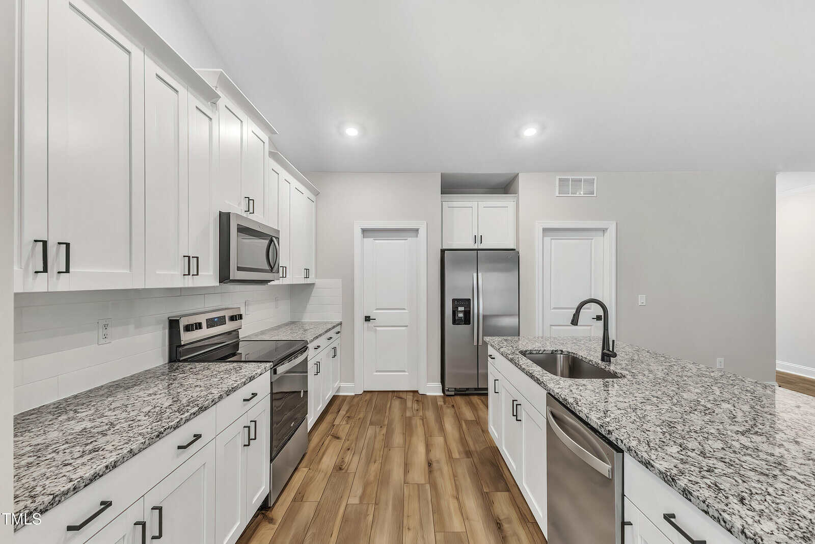Modern kitchen with white cabinets, stainless steel appliances, granite countertops, an island with a sink and dishwasher, wood-look flooring, and bright overhead lighting.