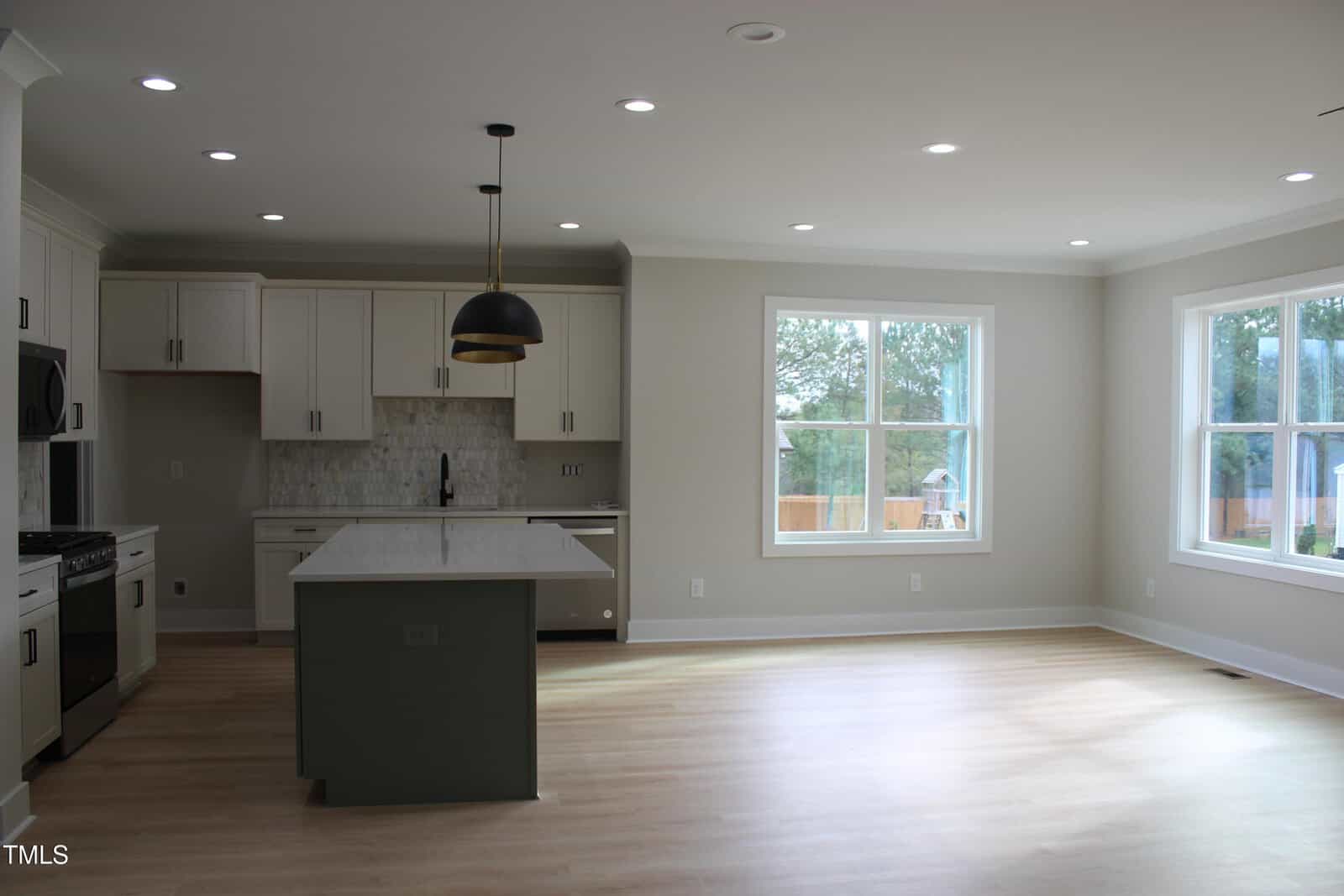 Modern kitchen with white cabinets, a center island, pendant lighting, light wood floors, and large windows letting in natural light. The space is open and unfurnished.