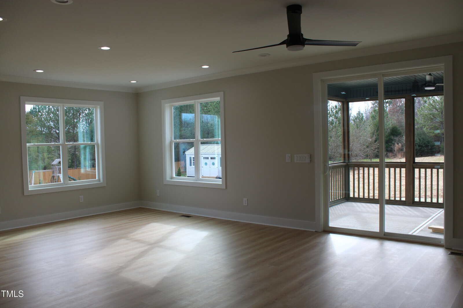 Bright, empty room with light wood floors, beige walls, two large windows, a ceiling fan, and sliding glass doors leading to a covered patio and grassy backyard.